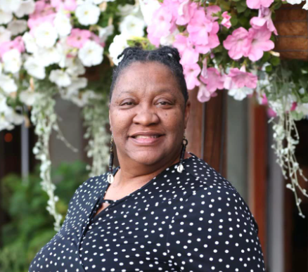 A smiling woman in a black polka dot top standing outdoors with pink and white flowers in the background.