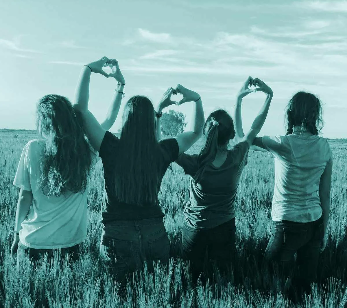 Four women standing with arms linked, forming heart shapes with their hands while looking out over a field, symbolising connection and community