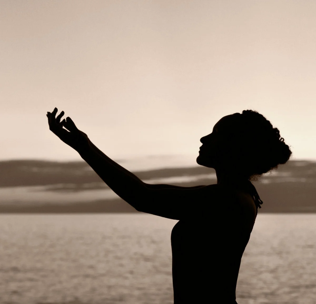 Silhouette of a woman raising cupped hands toward the sky by the ocean, symbolising empowerment, healing, and lifelong self-care