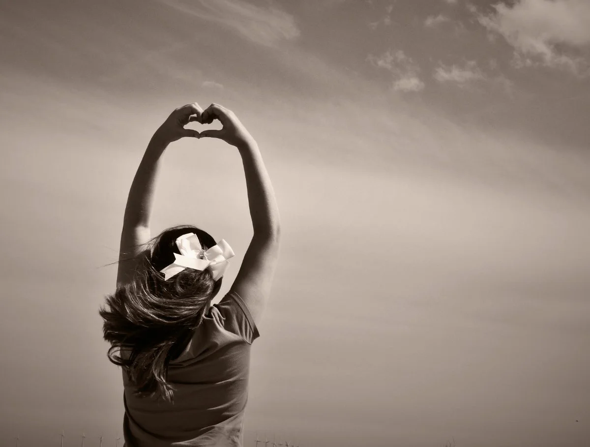 Woman seen from behind raising her hands to form a heart shape against the sky, symbolising self-love and personal growth