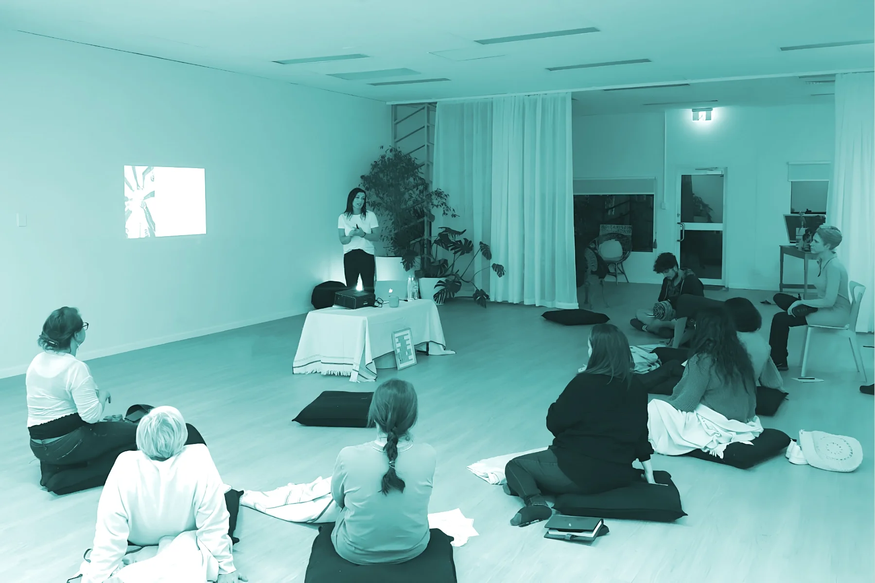 Kerry presenting in front of a group seated on the floor in a room with white walls and wooden floors.