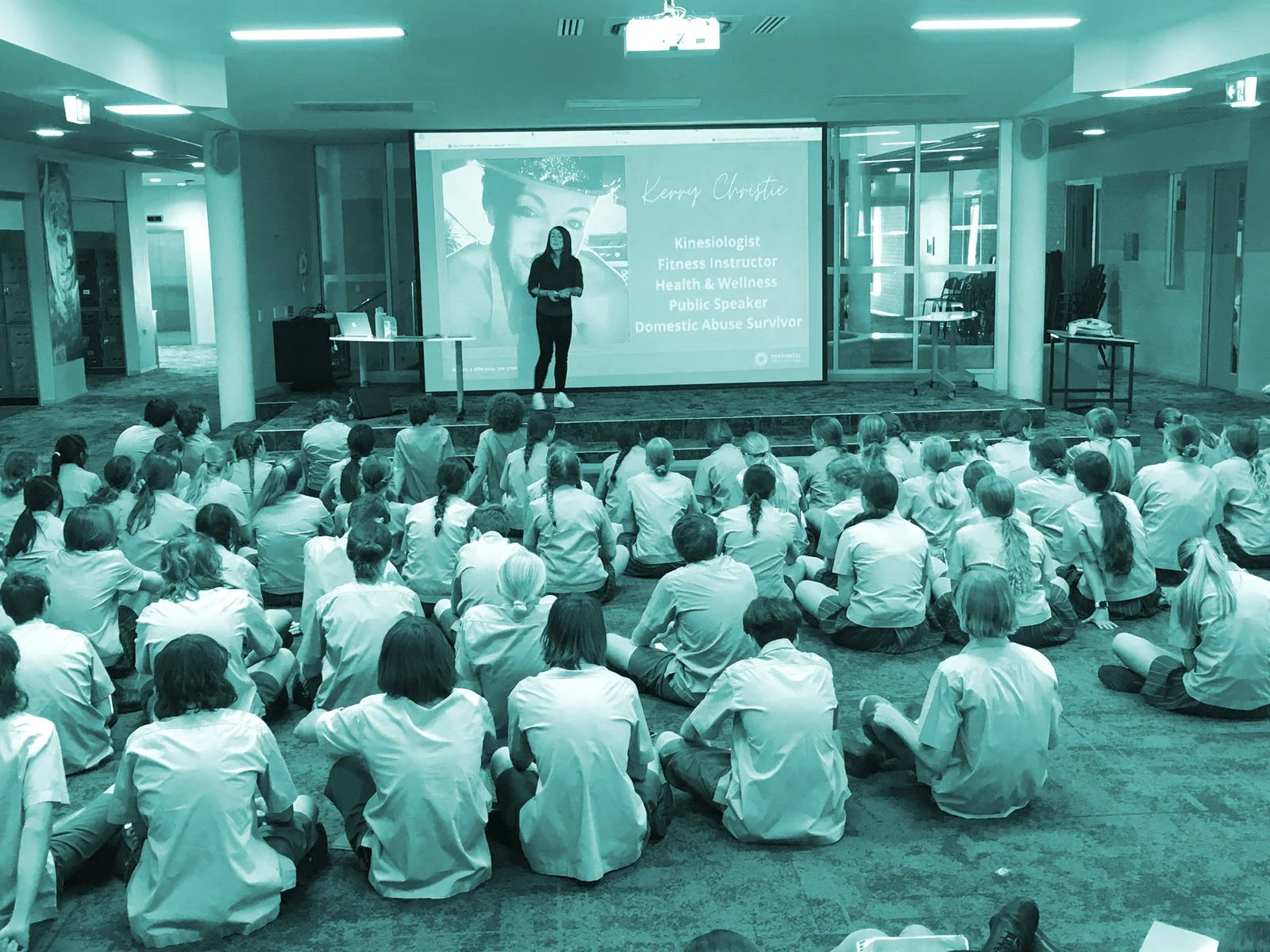 A woman giving a presentation to a group of students seated on the floor in a large room. The presentation slide shows her name, Kerryn Christie, with titles such as Kinesiologist, Fitness Instructor, Health & Wellness Public Speaker, Domestic Abuse Survivor.