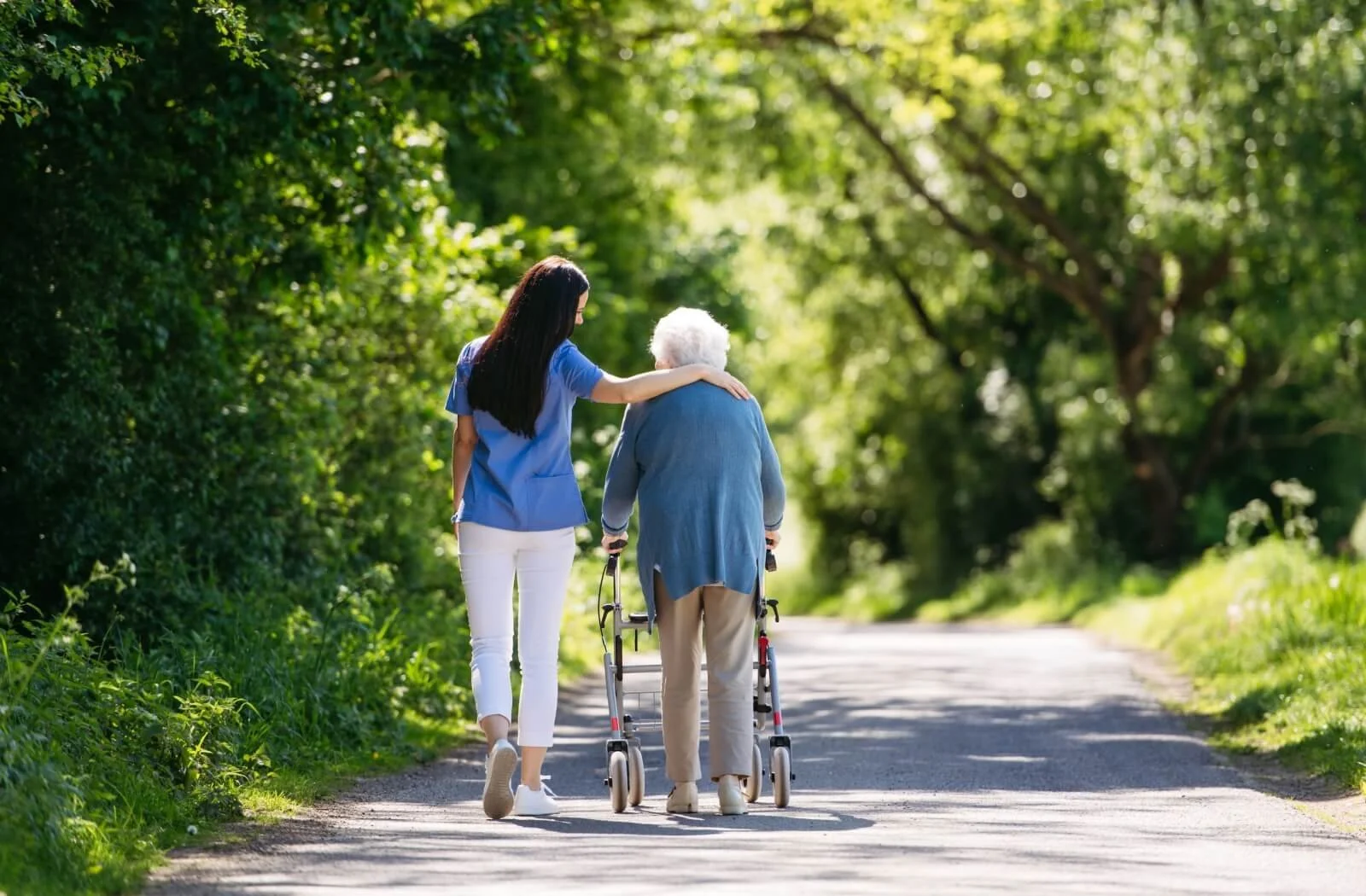A young woman in blue scrubs supporting an elderly woman with white hair walking with a walker on a tree-lined path.