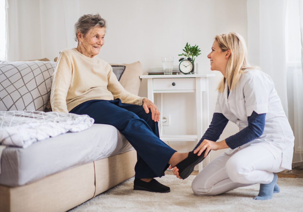 A caregiver helping an elderly woman put on her shoe in a cozy, well-lit bedroom.