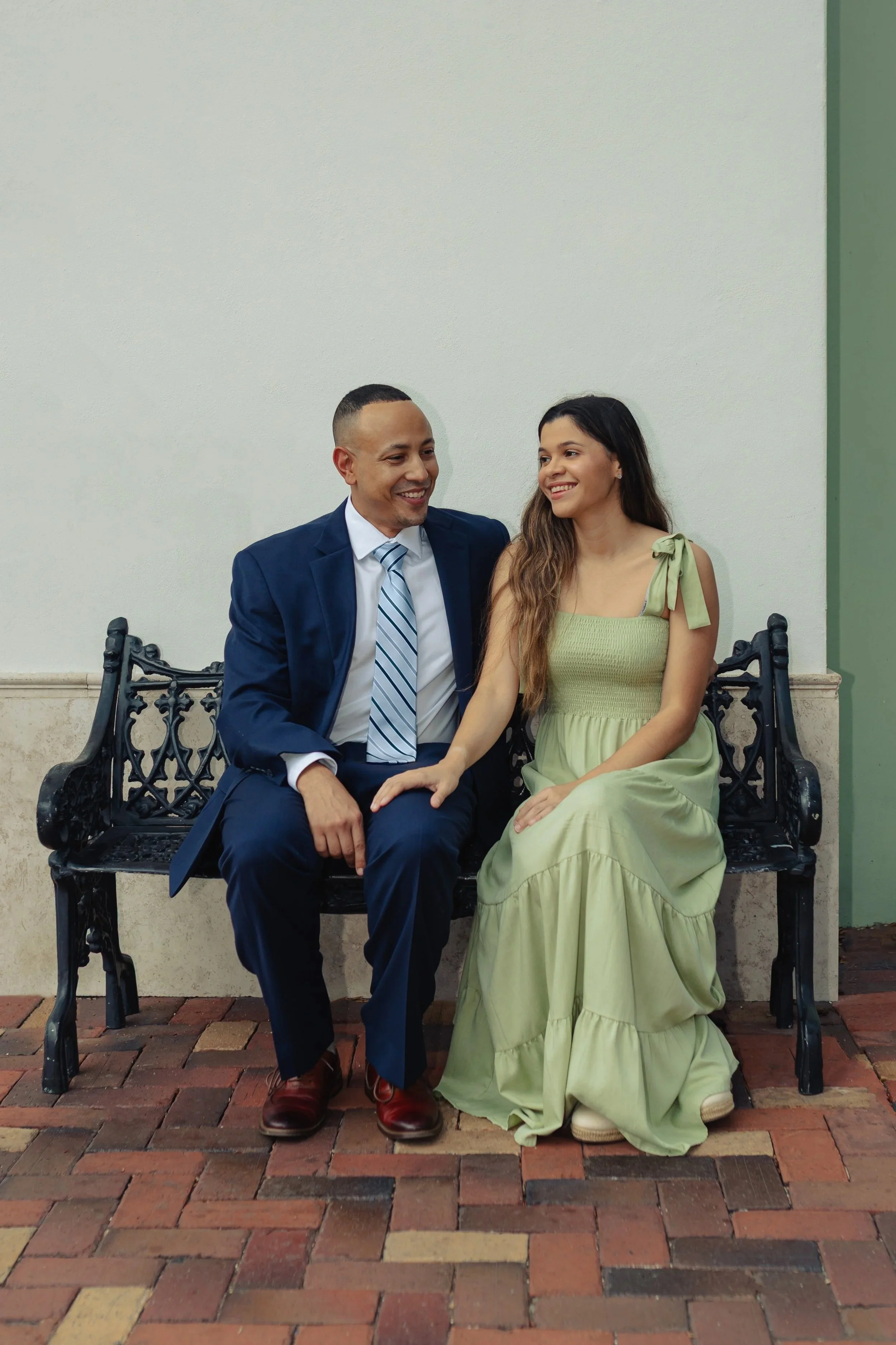 Jonathan Harris in a blue suit and his wife Jessica in a green dress sitting on a black ornate bench, smiling and looking at each other, outdoors on brick pavement.