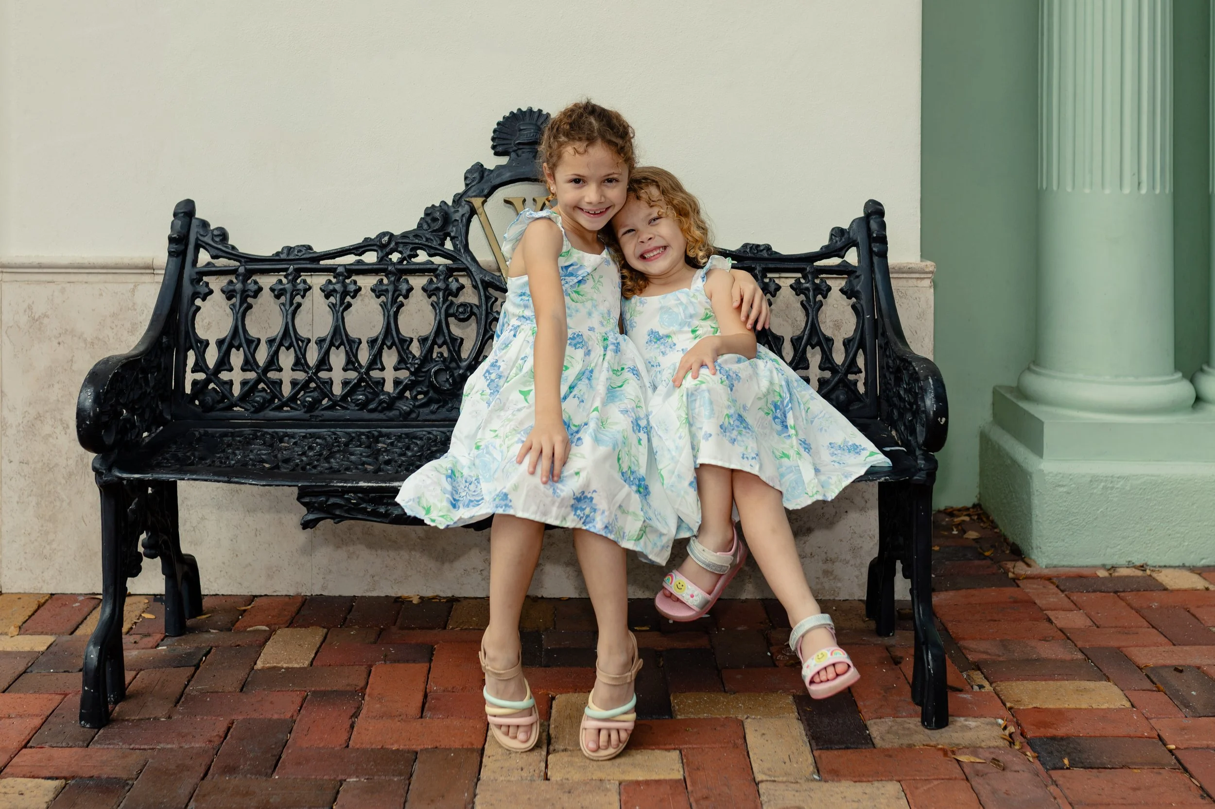Jonathan Harris' Daughters in matching floral dresses smiling on a black ornate park bench on brick pavement.