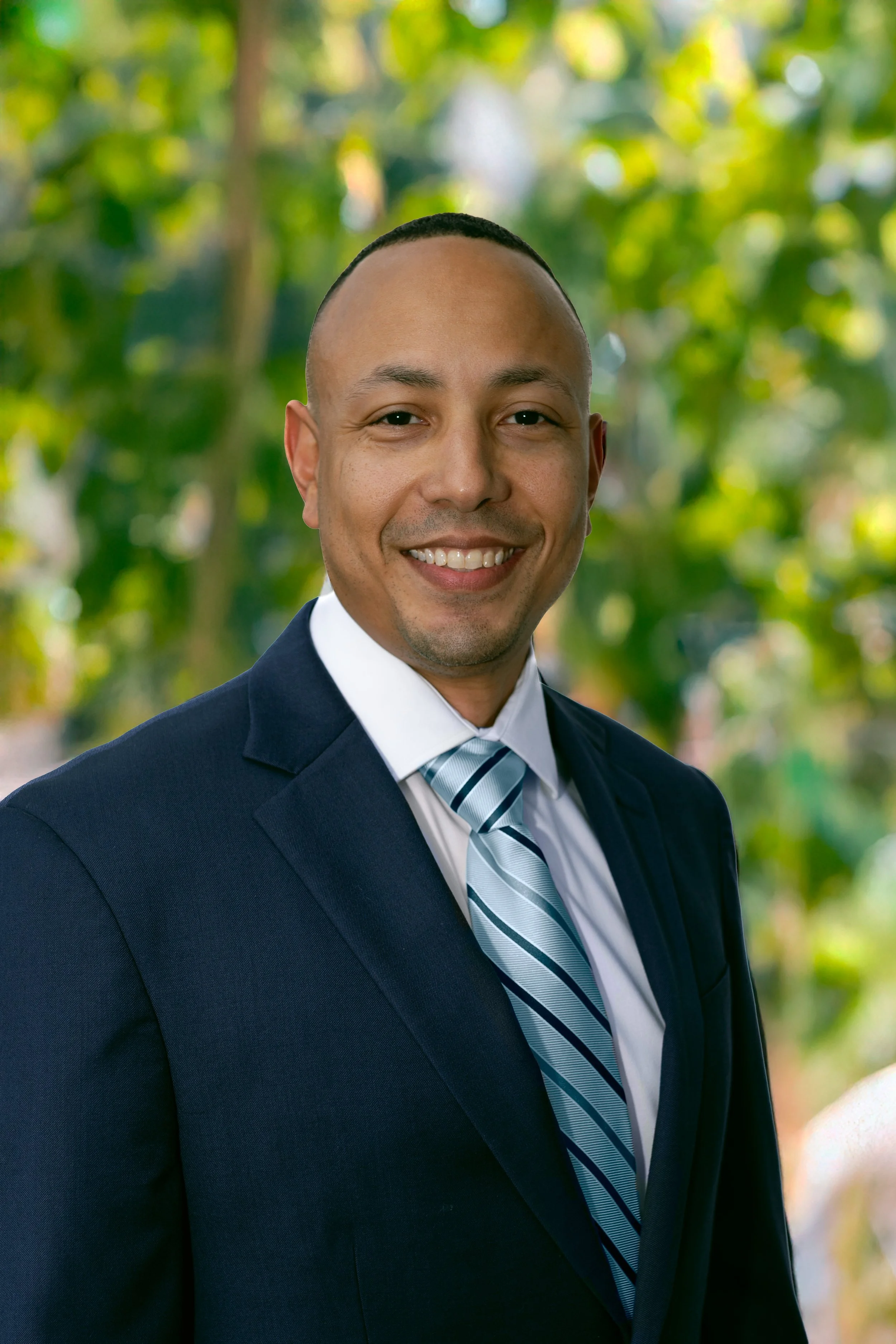 Jonathan Harris, candidate for Florida's 16th congressional district, in a navy suit, white shirt, and light blue striped tie stands outdoors with green foliage in the background.