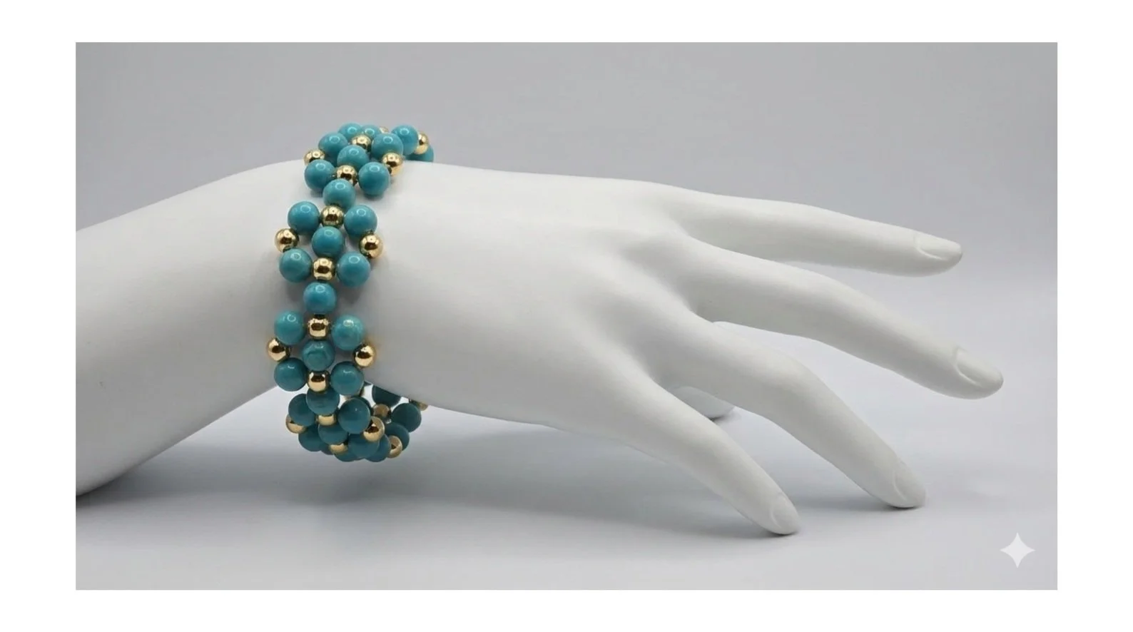 A white mannequin hand wearing a blue beaded bracelet with gold accents, resting on a plain white surface against a gray background.