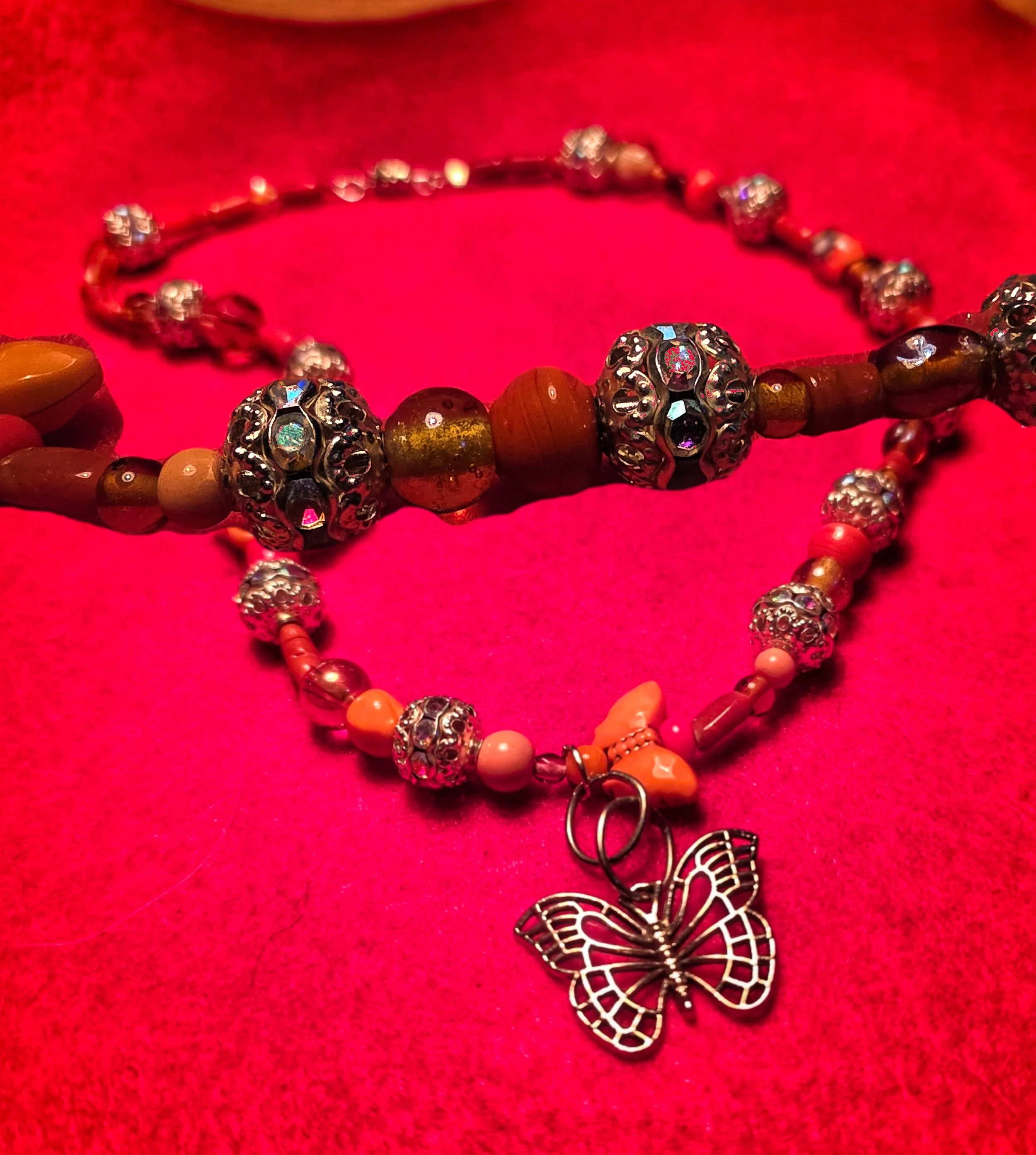 Close-up of a pink beaded necklace with silver decorative beads and a butterfly pendant, laid on a pink fabric background.
