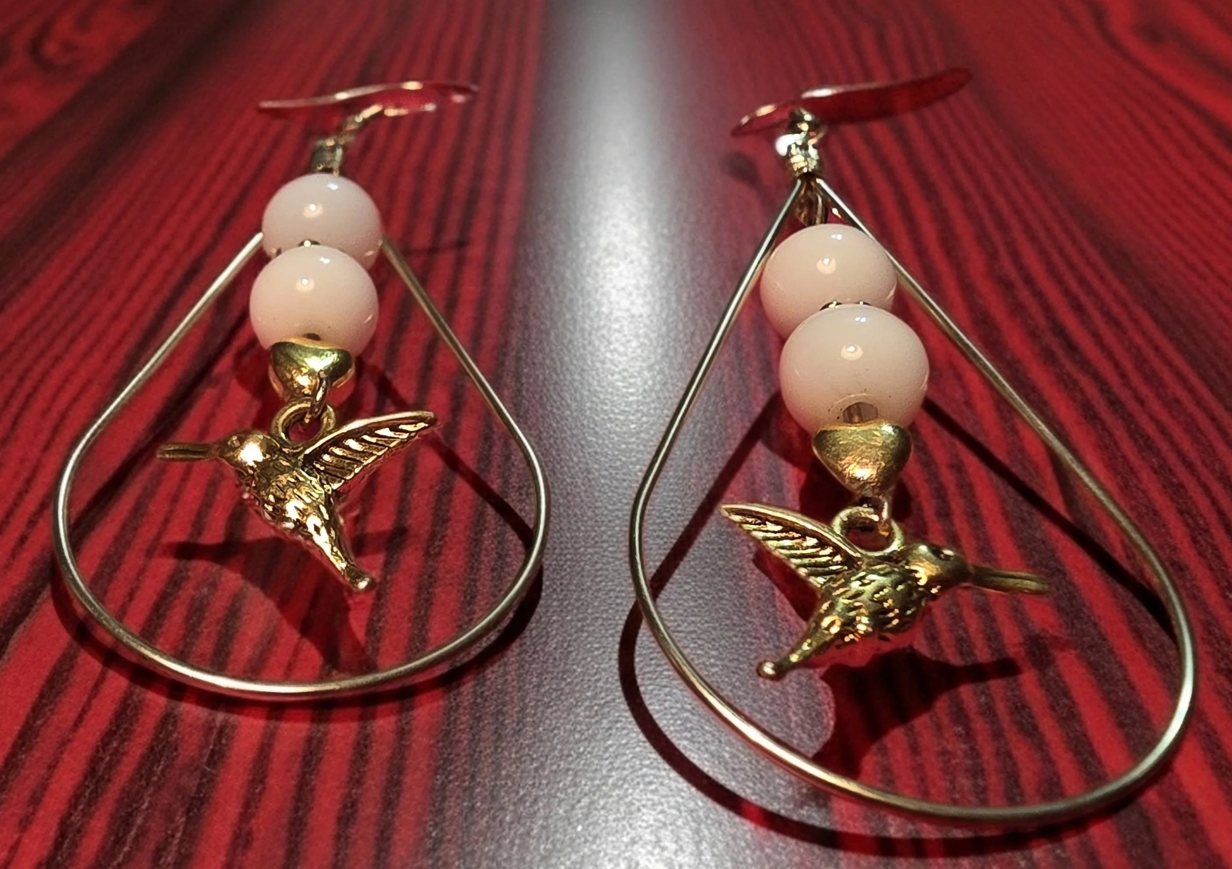 Pair of gold and pink hummingbird earrings with pink beads on a dark striped wooden surface.