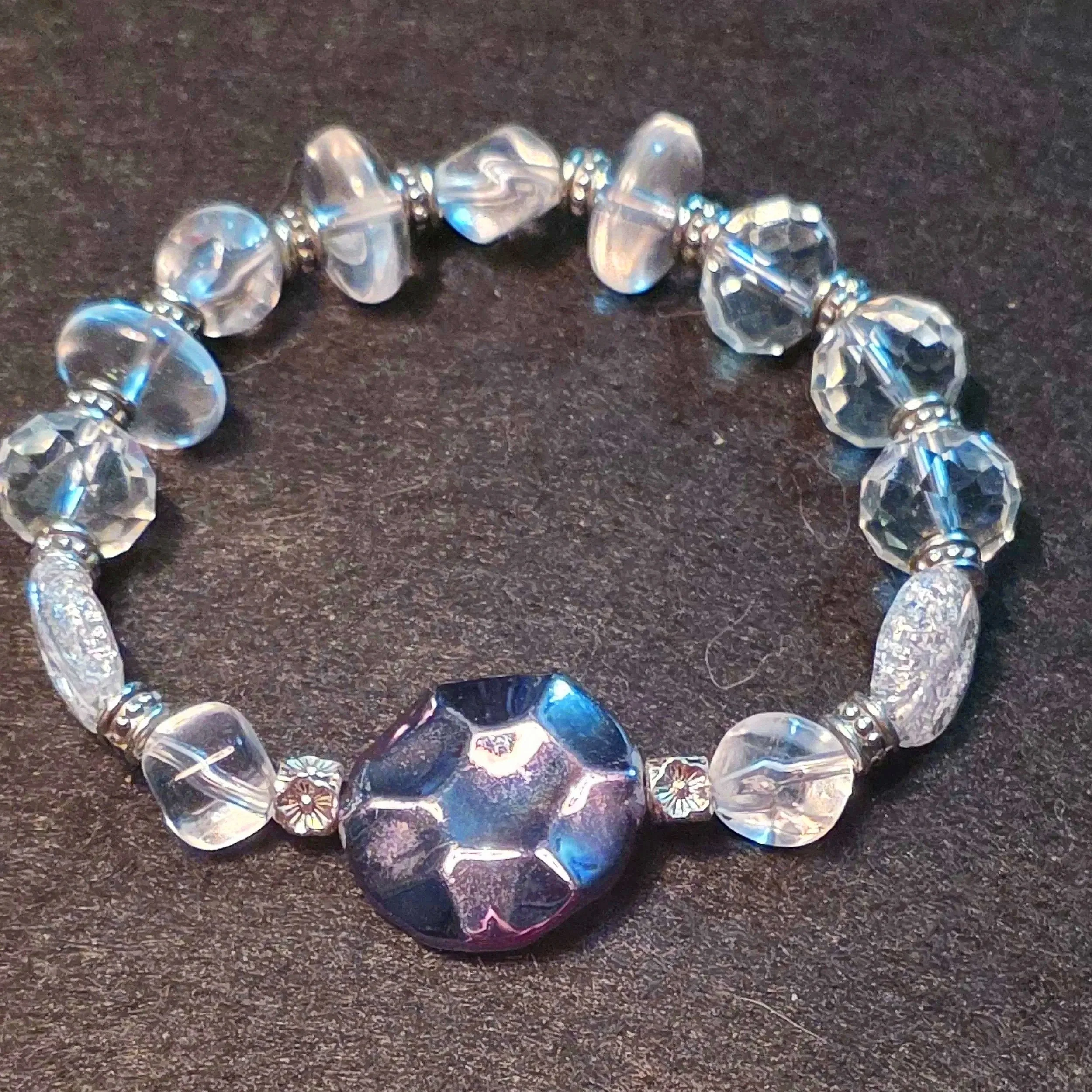 A beaded bracelet made of clear and iridescent crystals with a large central bead on a dark surface.
