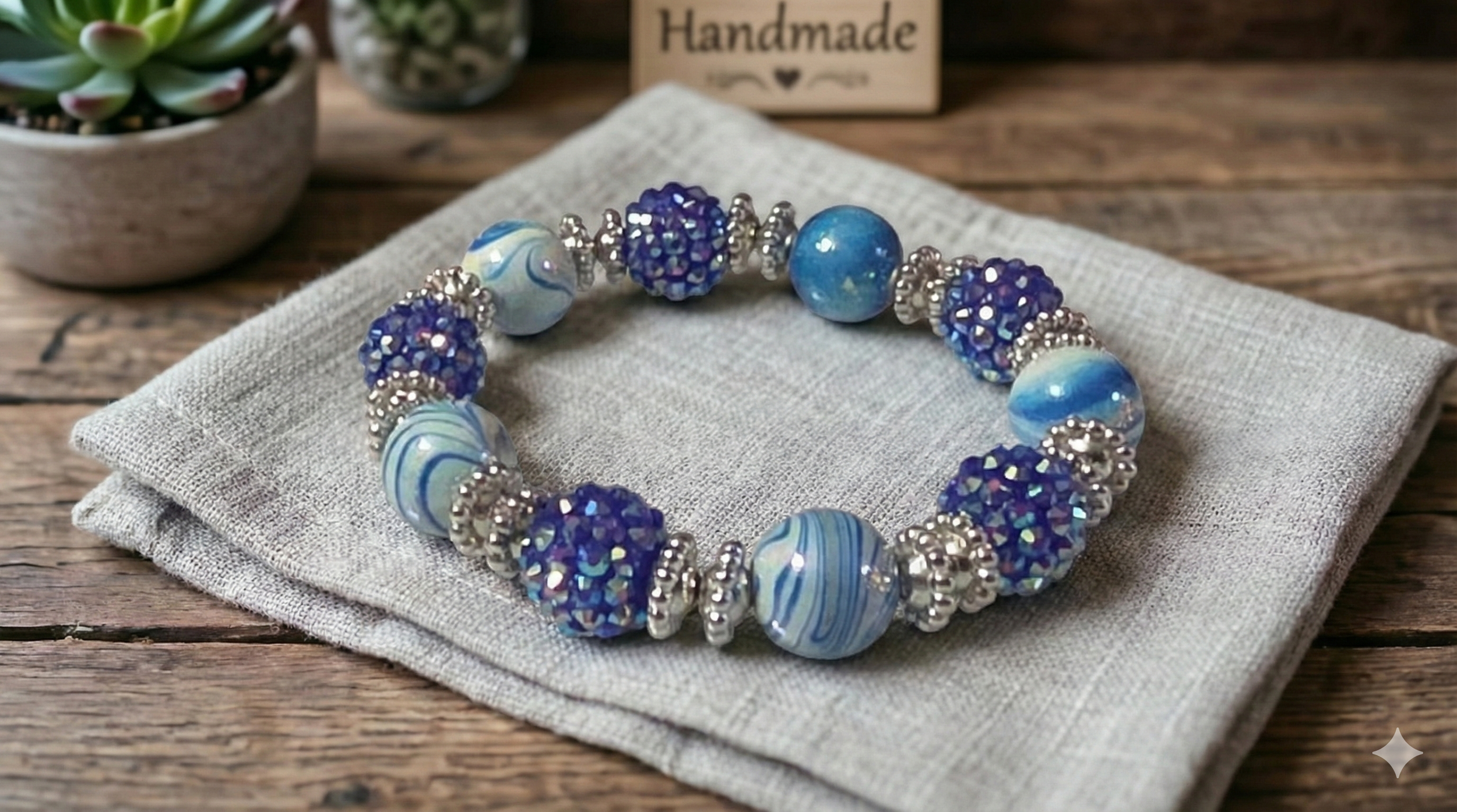 Handmade blue and purple beaded bracelet with silver accents, placed on a beige fabric cloth on a wooden surface, with a small succulent plant and a 'Handmade' sign in the background.