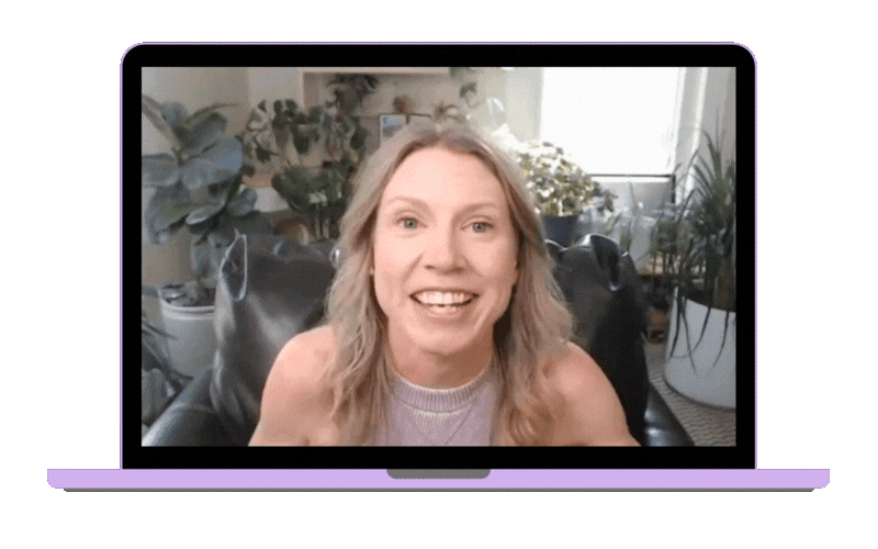 A woman with blonde hair smiling on a video call, sitting on a black leather chair in a room with plants and natural light.