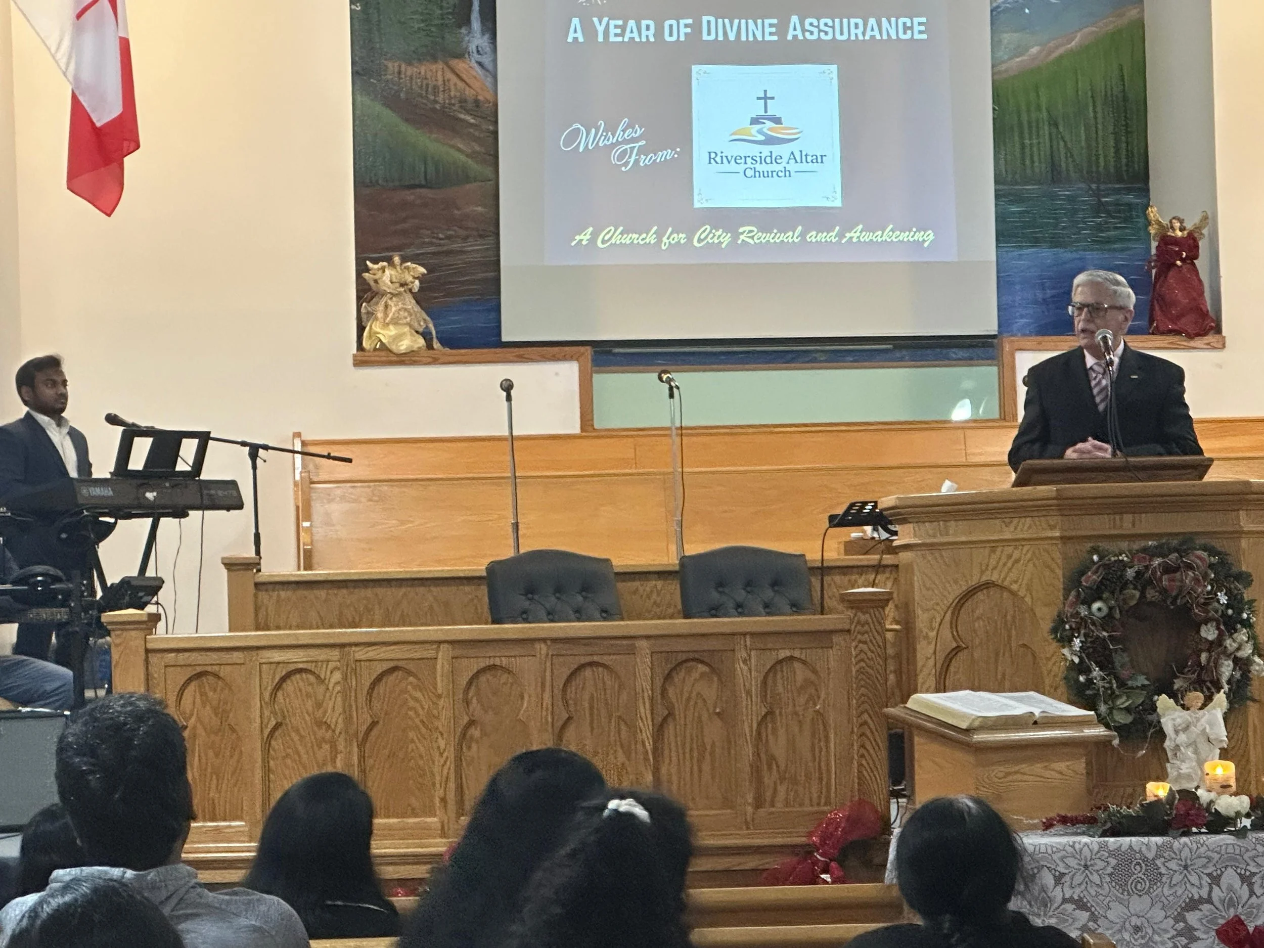 A man in a suit speaking at a church podium decorated with a Christmas wreath during a church service. A keyboard player is on the left side of the stage. A large projected slide behind him reads: "A Year of Divine Assurance, Wishes From: Riverside A