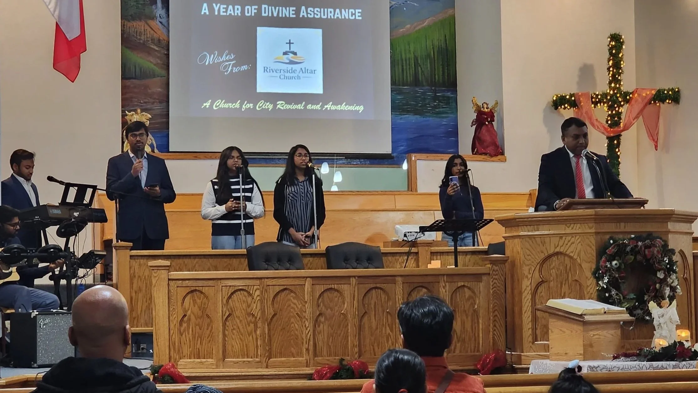 A church service with a male speaker at the pulpit, five people standing and singing on the stage, and a band to the left. The backdrop displays the message, 'A Year of Divine Assurance' and wishes from Riverside Altar Church. Christmas decorations, 