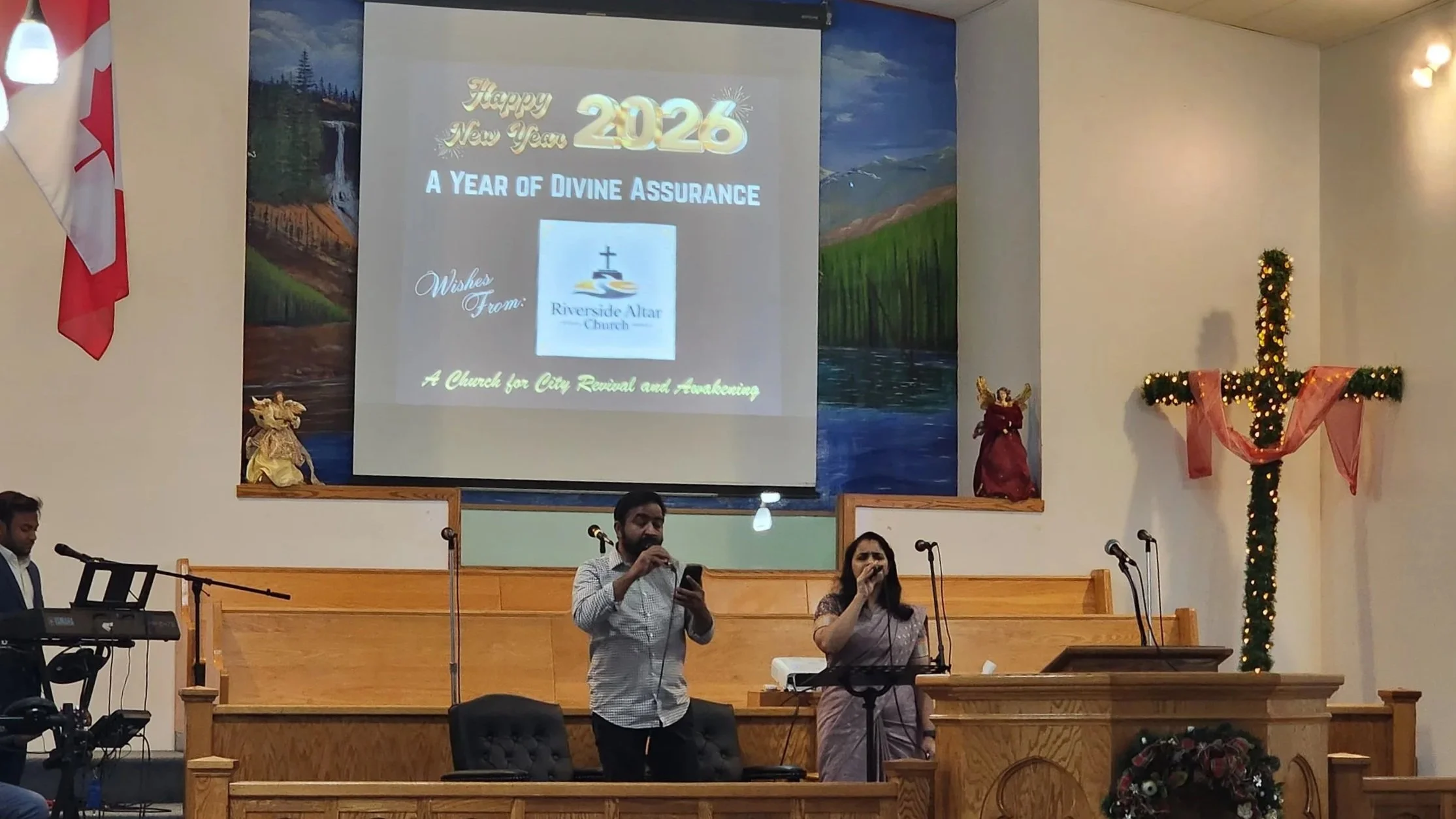 A church stage decorated for Christmas with a large cross wrapped in pink ribbon and lights on the right, a screen displaying a New Year 2026 message from Riverside Altar Church on the center, and two people singing into microphones in front of the s