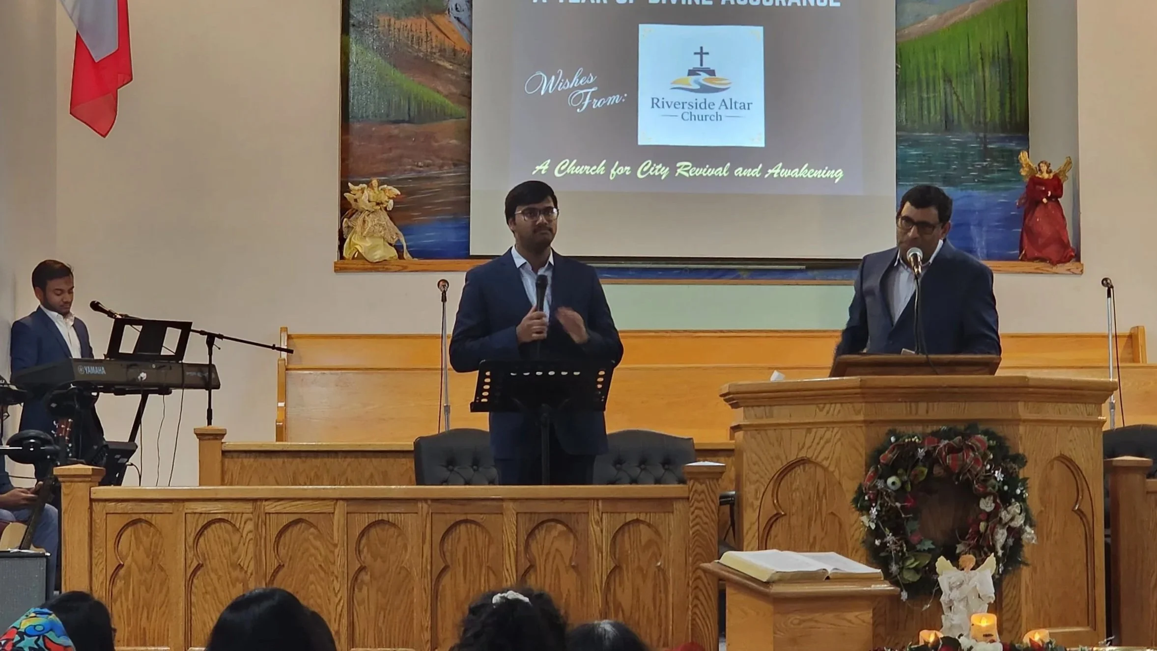Church service inside a church with two men speaking at the pulpit, one playing keyboard and another on the podium, with a large screen behind displaying a church logo and a message. The church is decorated with a wreath and candles.