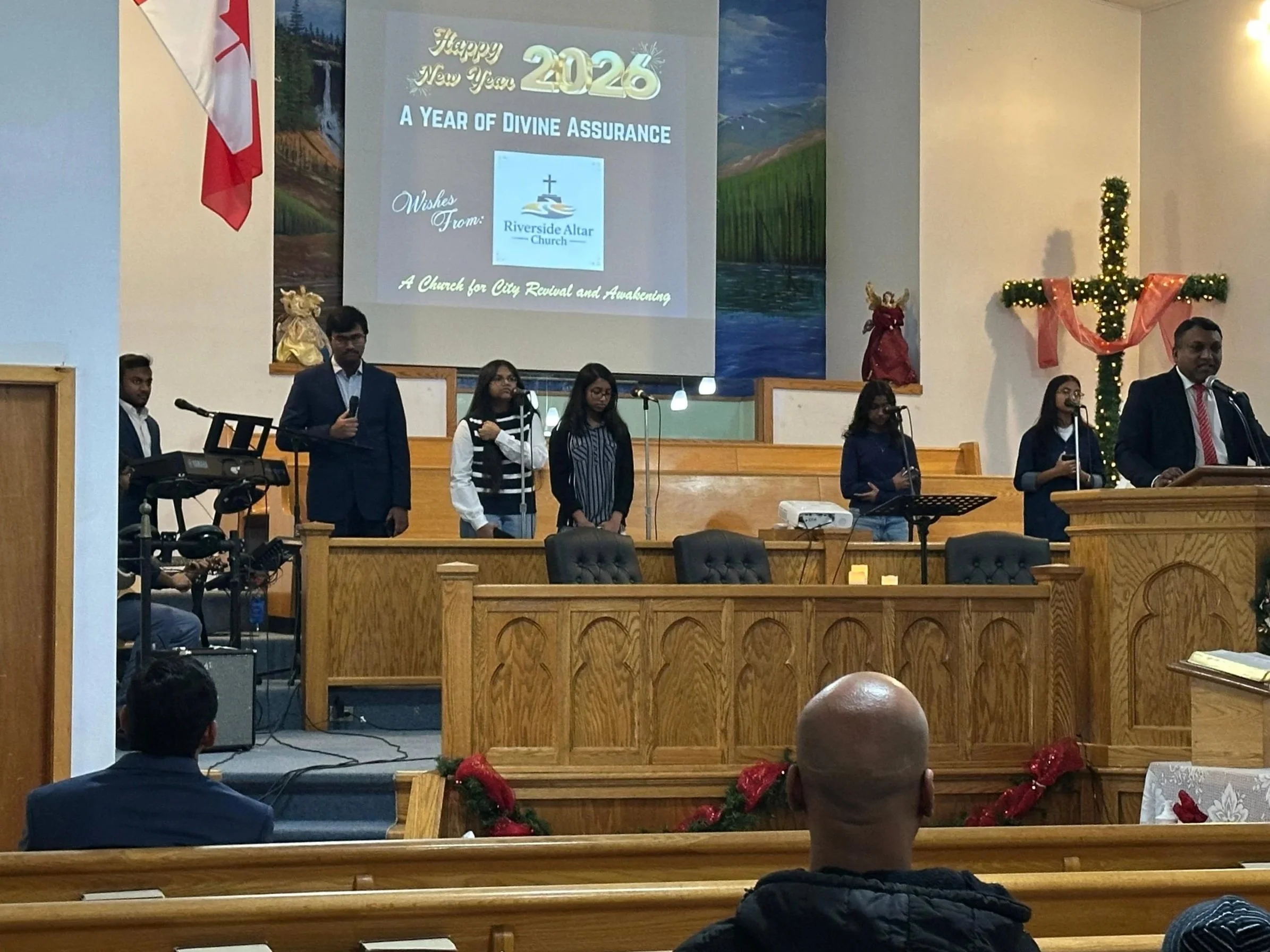 Church service with people standing on stage, singing or speaking into microphones, led by a man at a wooden pulpit. A large Christmas cross decorated with Christmas lights and ribbons is on the wall behind them. A screen displays a New Year message 