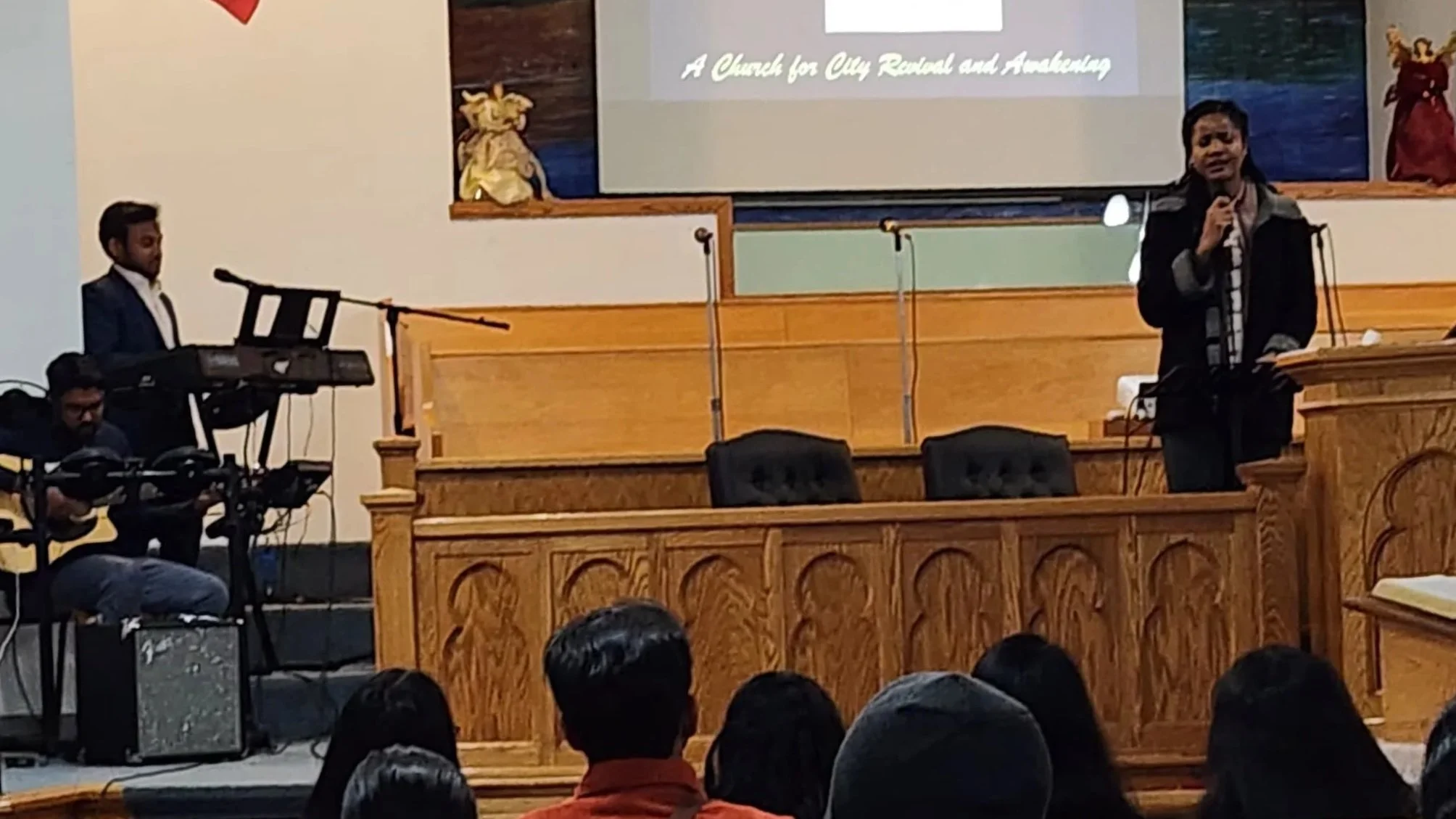 A woman is speaking at a podium during a church service or event, with a man playing keyboard and another person sitting nearby. There are several people in the audience, and a large screen in the background with text that says 'A Church for City Ren