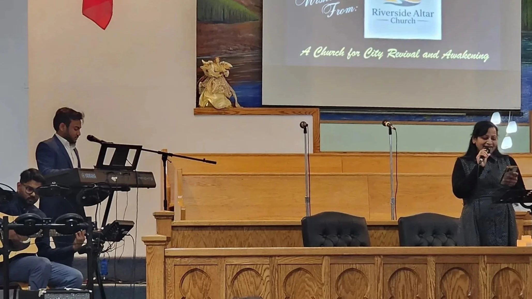 A group of people on a stage during a church service, featuring a woman singing into a microphone, a man playing keyboard, and another person seated with headphones. A large screen behind displays a message from Riverside Altar Church, promoting city