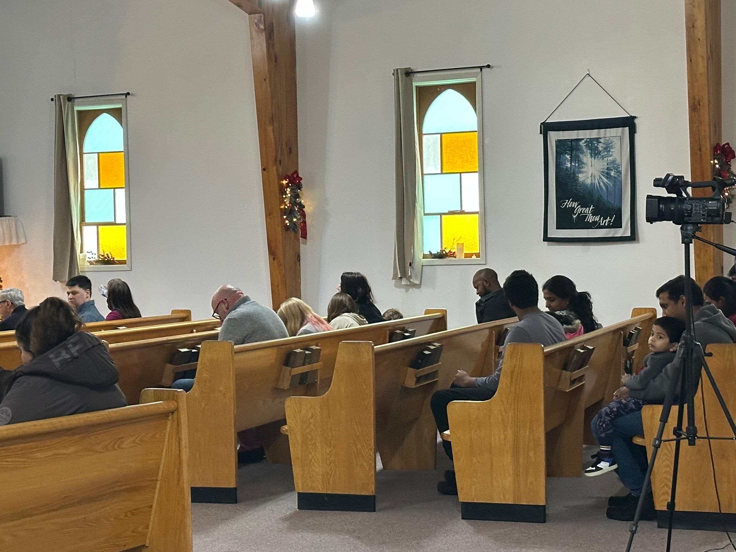 People seated in wooden church pews inside a church during a service or event, with three stained glass windows on the wall, decorated for the holidays, and a video camera on a tripod to the right.