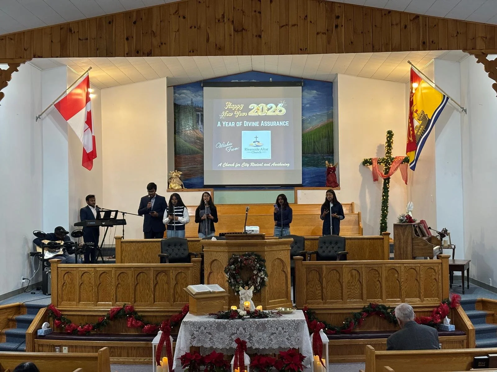 Church congregation celebrating New Year's 2026 with a decorated altar, gospel musicians, and a holiday-themed backdrop featuring flags and Christmas decorations.