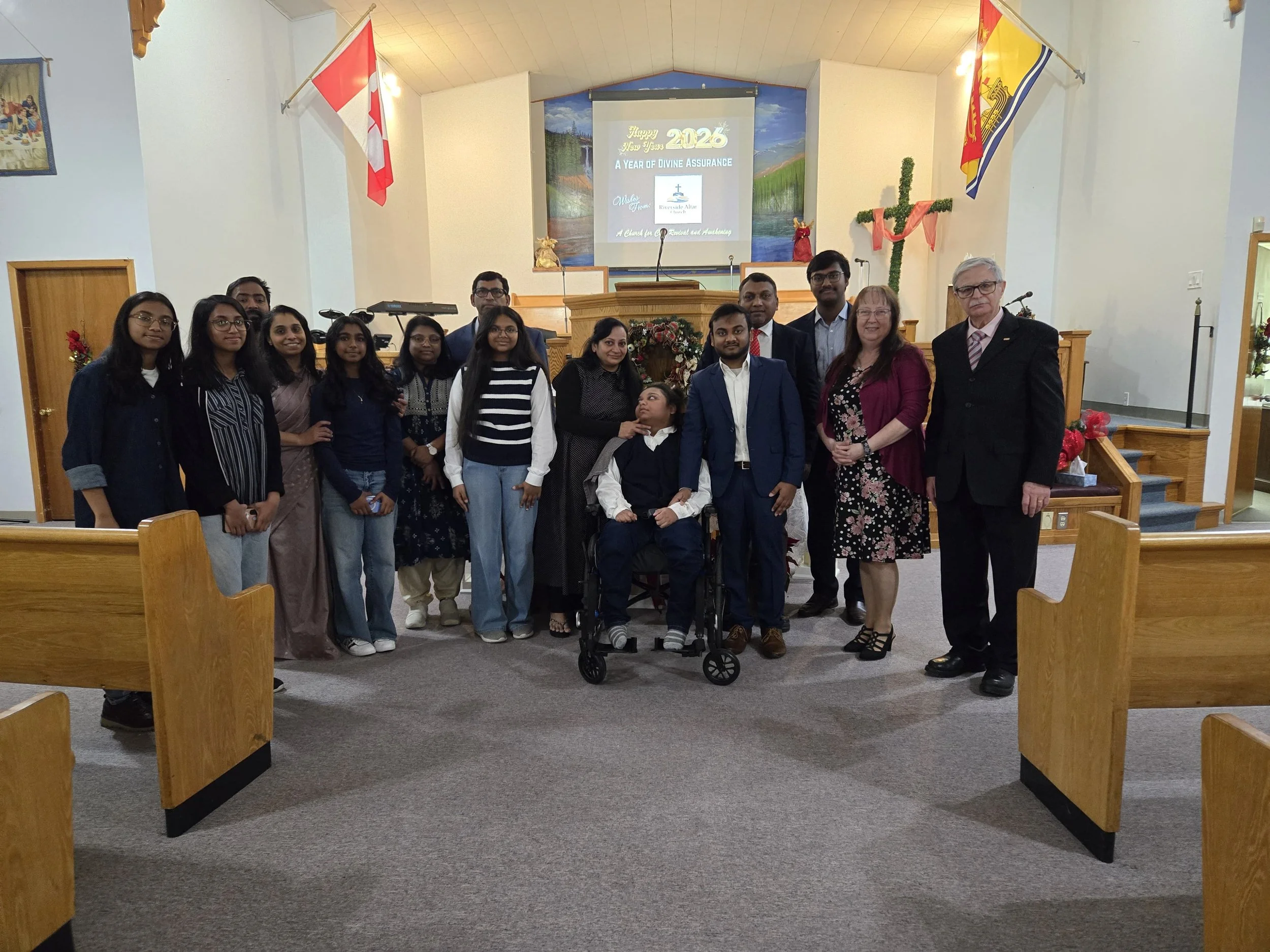 A group of people gathered inside a church, standing in front of the altar. The group includes men, women, and children, with some dressed in formal attire. A presentation screen behind them displays a New Year message for 2026, symbolizing hope and 