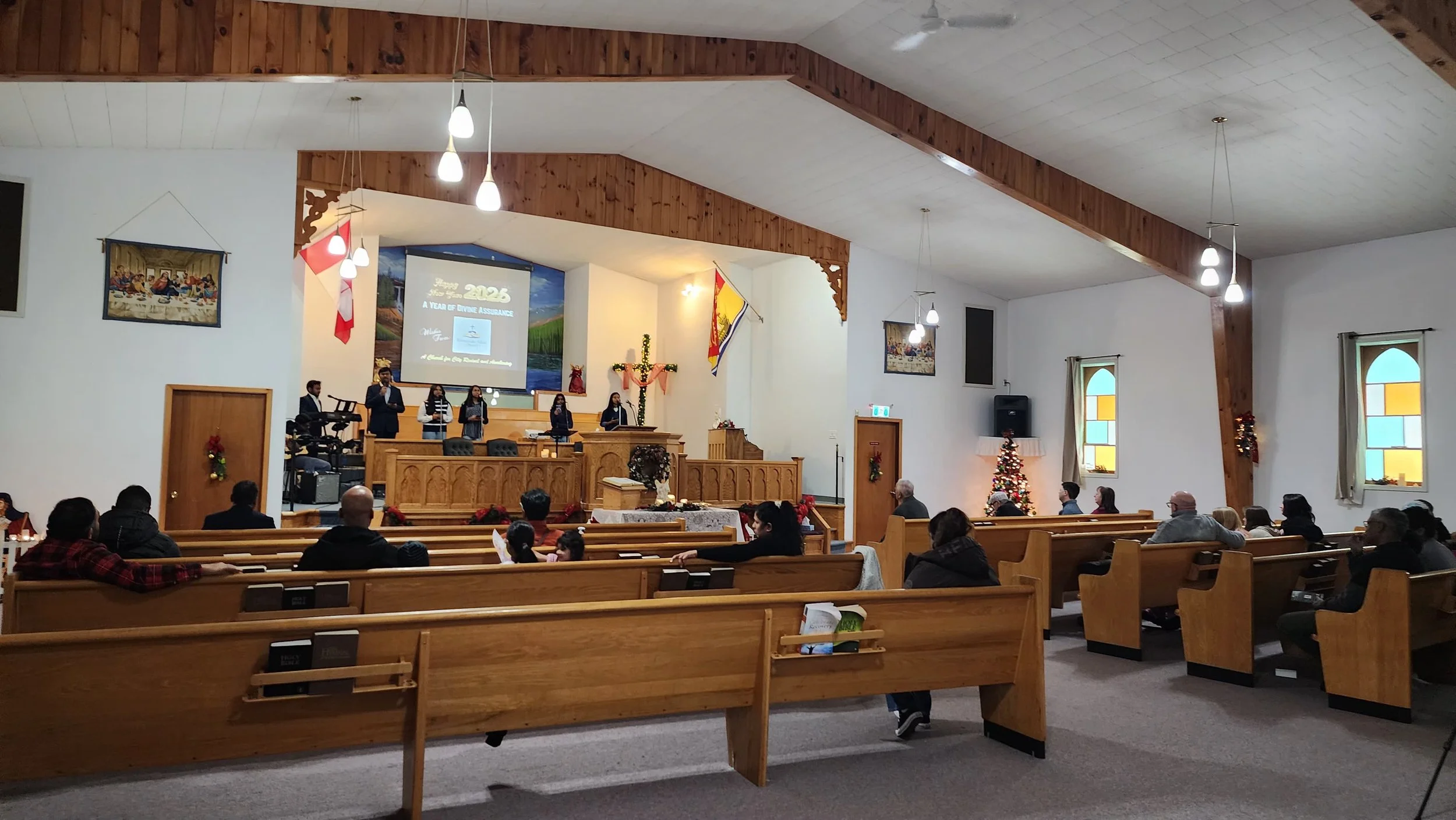 People seated in pews inside a church, listening to a band and choir on stage, with Christmas decorations and a Christmas tree visible.