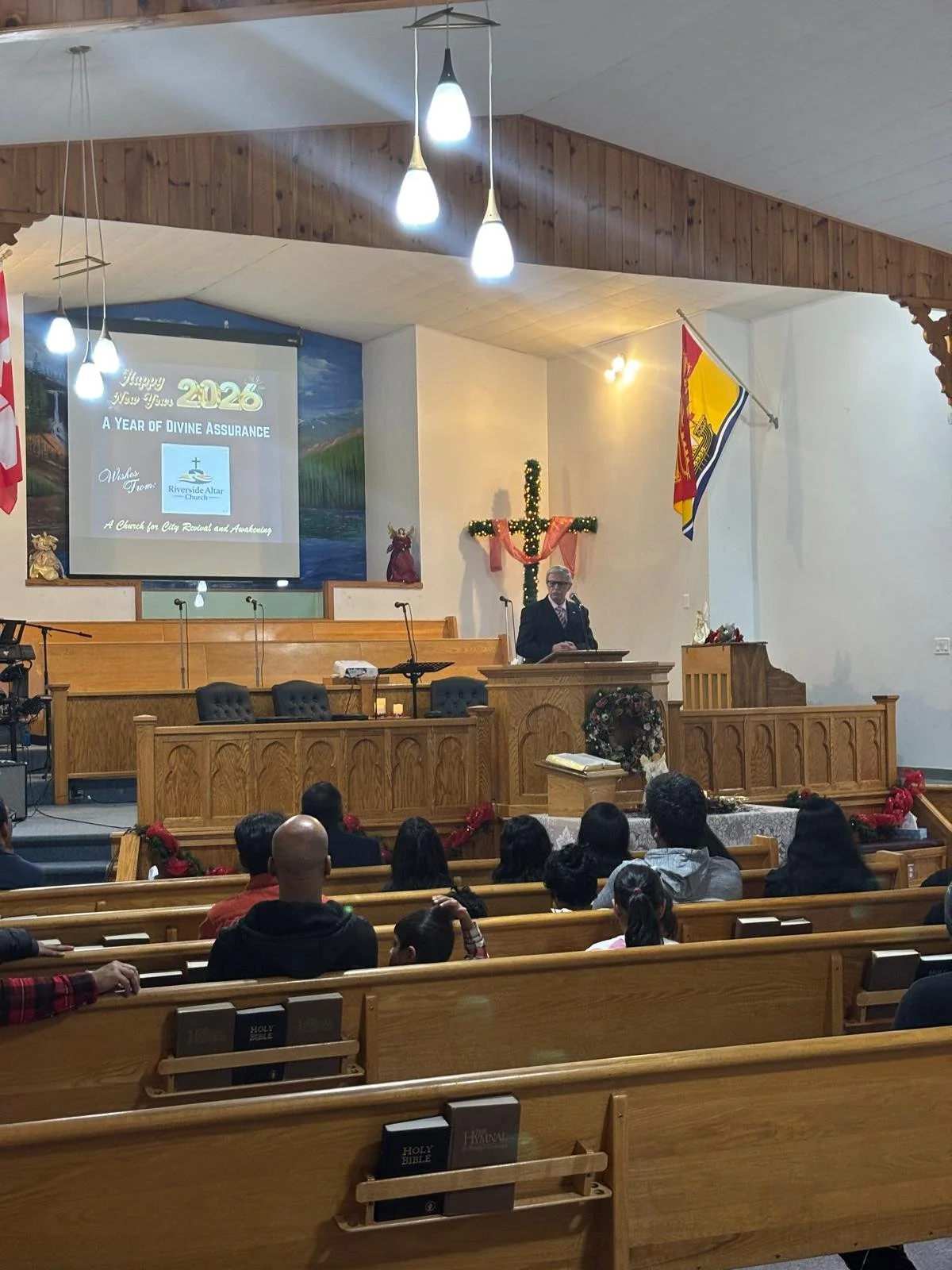 A church service inside a church with a congregation, a pastor at the pulpit, and Christmas decorations including a cross with a garland and a wreath. The screen displays a message for 2026 and a church logo, with flags and lighting fixtures overhead