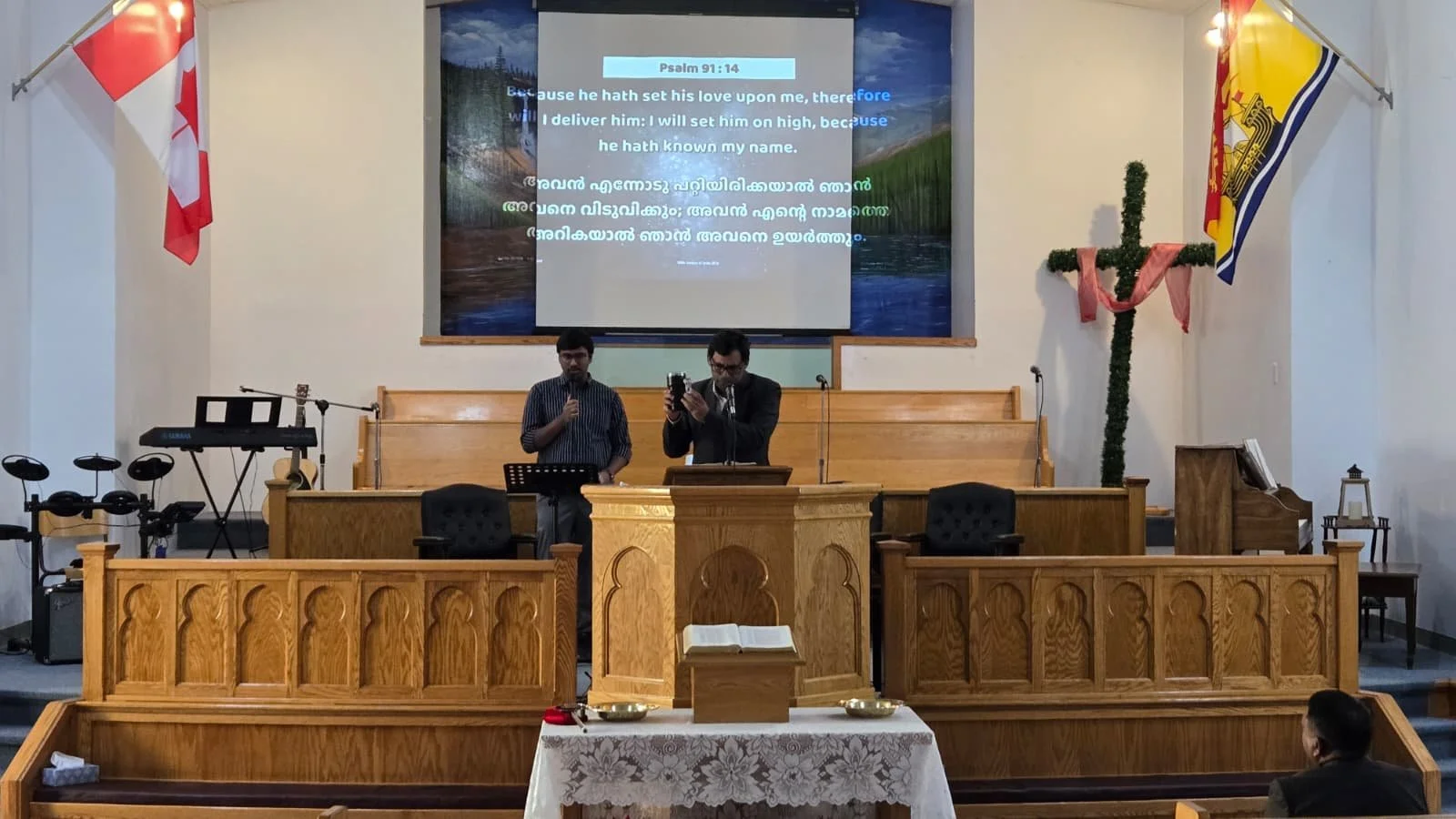 Inside a church with wooden pulpit and music instruments, two men singing or speaking at the front, a large cross decorated with pink cloth, flags on either side, and a screen displaying Bible verses in English and another language.