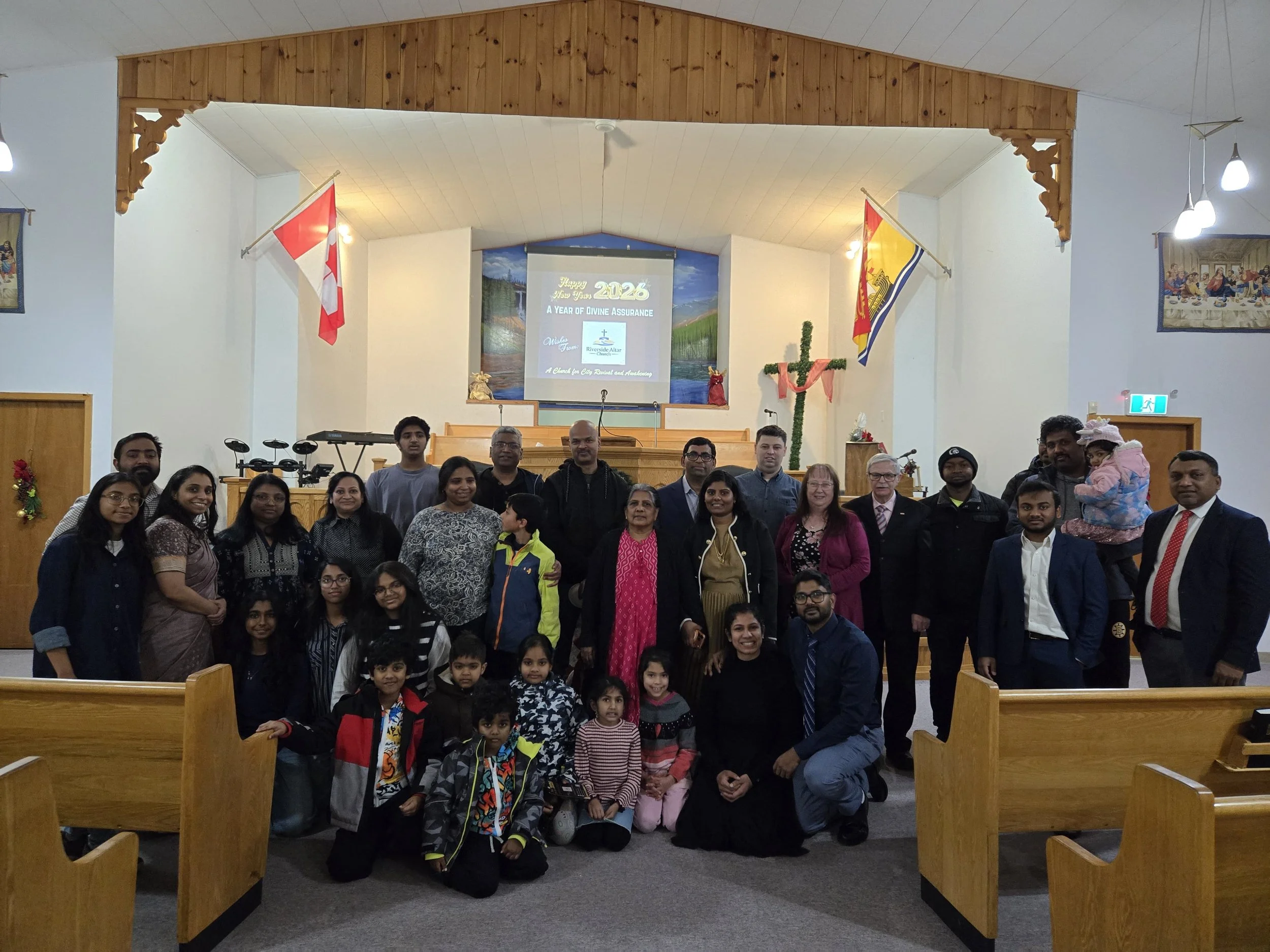 A diverse group of people, including adults and children, gathered in a church sanctuary for a group photo. The background features a cross decorated with flowers, flags, and a church altar. A digital screen displays a New Year message for 2026.