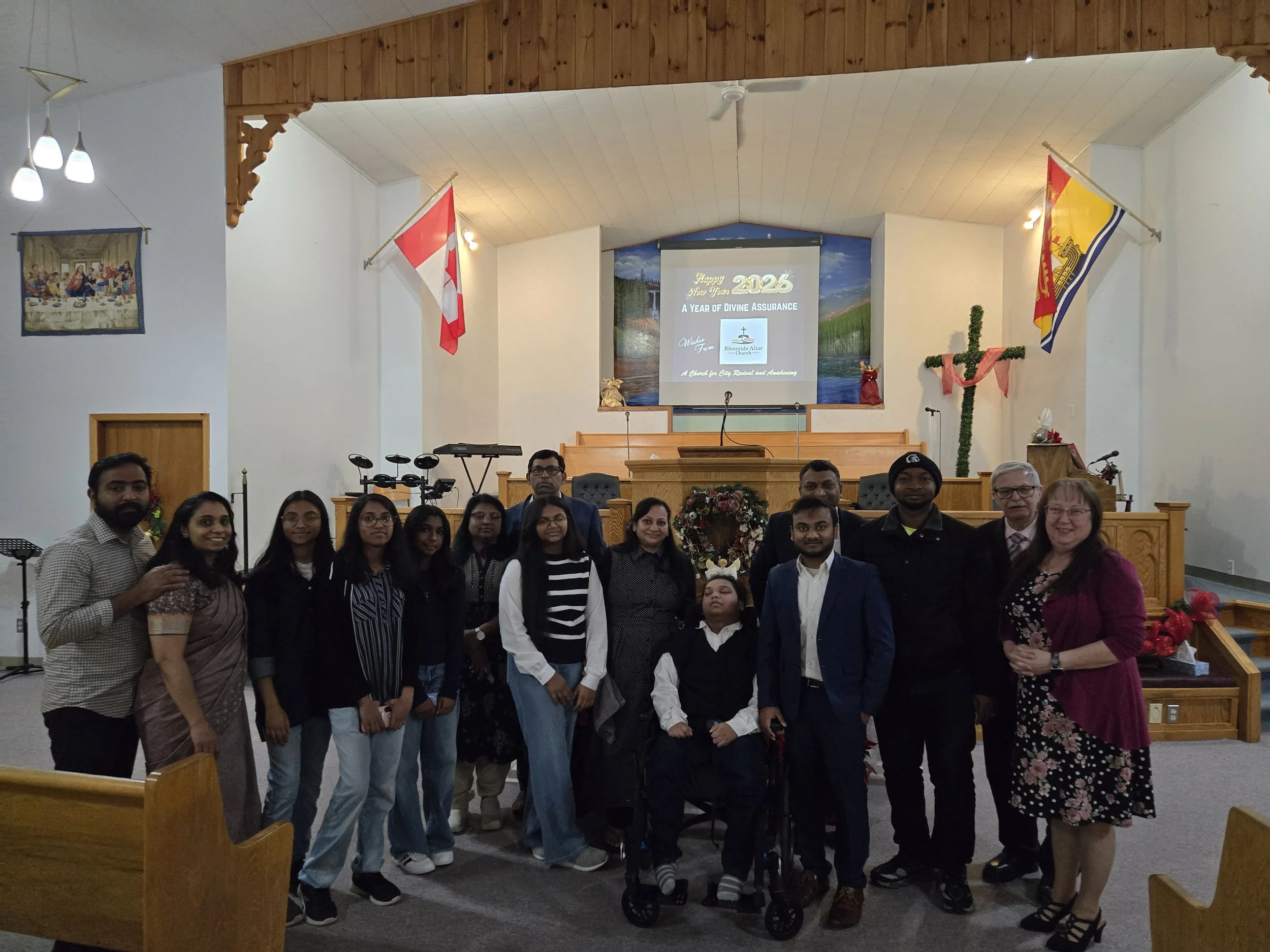 Group of people in a church, standing and sitting in front of the altar. The church has flags, musical instruments, and a screen displaying a New Year's message for 2026.