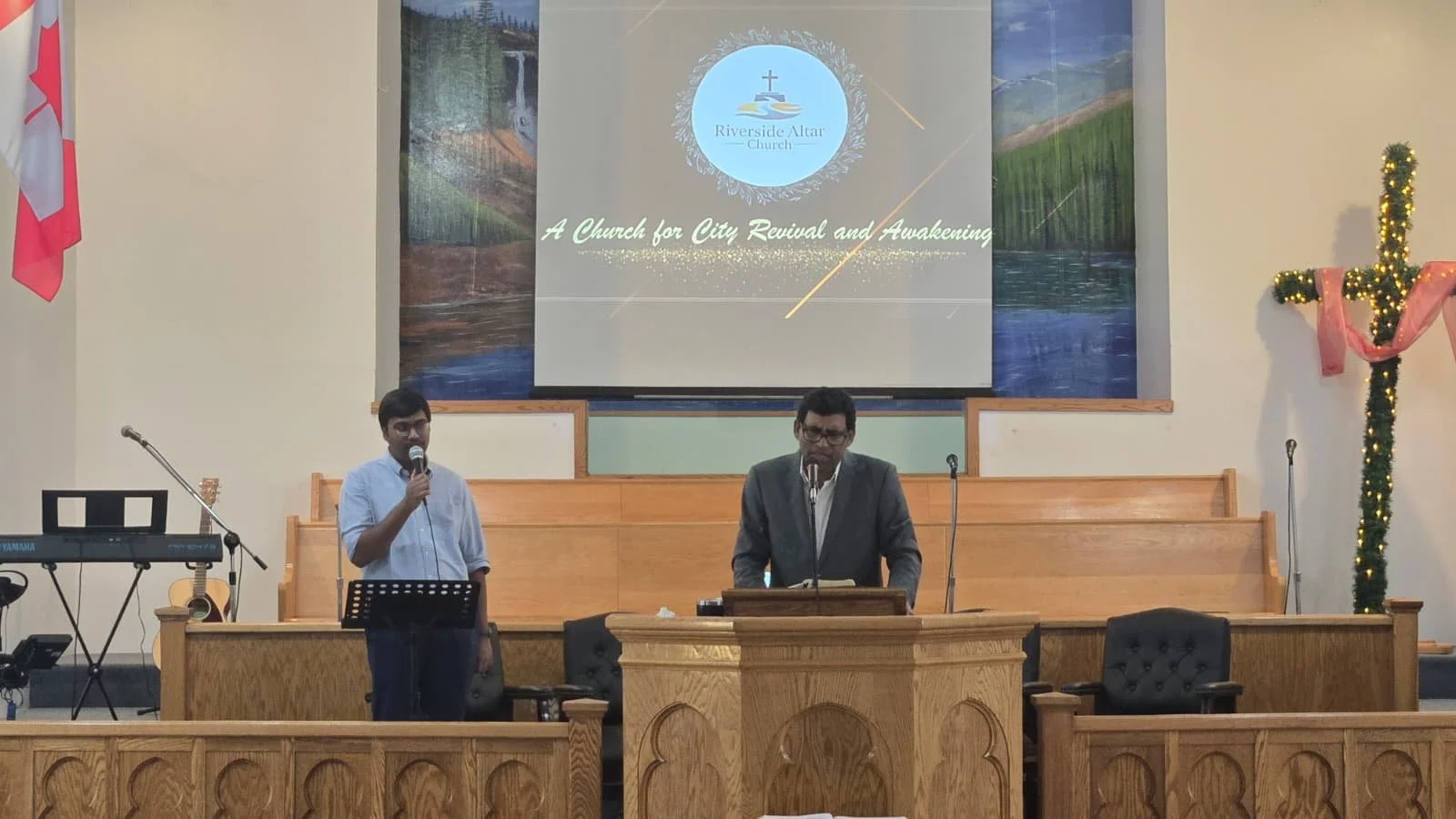 Two men standing at the pulpit in a church, one speaking into a microphone and the other reading from a book, with a large screen behind them displaying a church logo and the words 'A Church for City Revival and Awakening'. There is Christmas decorat