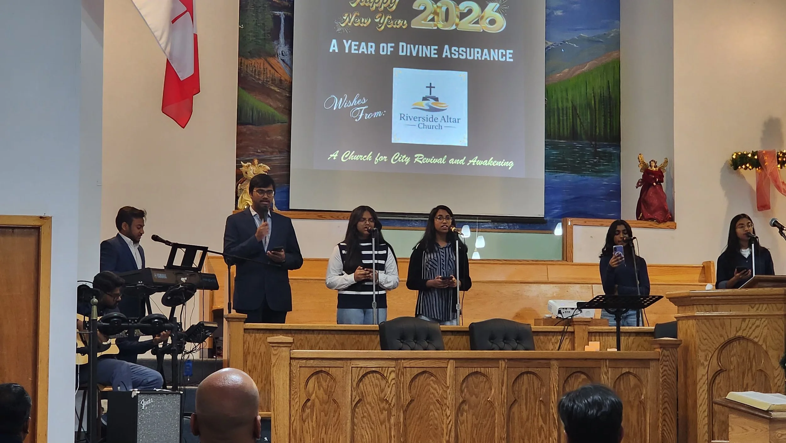 Group of six people singing or speaking into microphones on stage at Riverside Altar Church, with a large screen behind them displaying a New Year message for 2026 and wishes from the church. The stage has wooden paneling, and there are religious sta