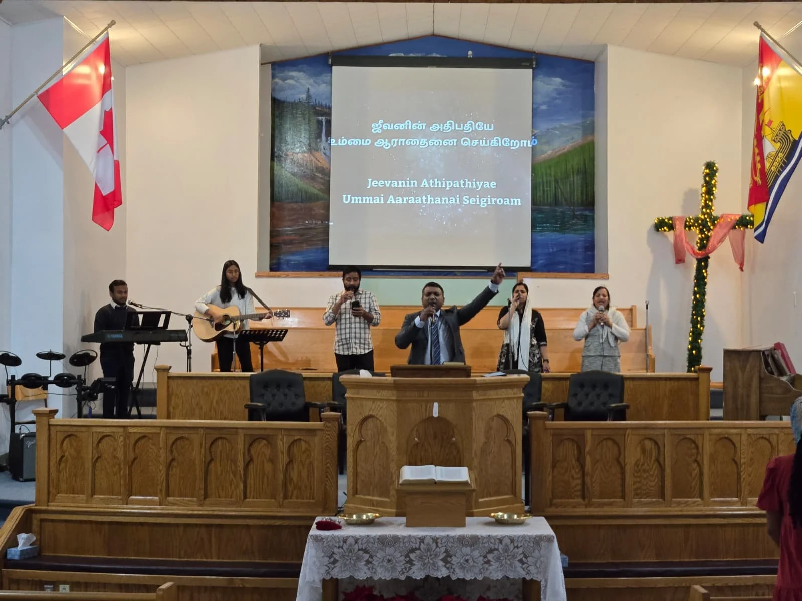 A church service with musical performers, including a guitarist, keyboard player, singers, and a preacher, on the stage. The stage has a screen with text and is decorated with national flags and Christmas decorations, including a cross and Christmas 