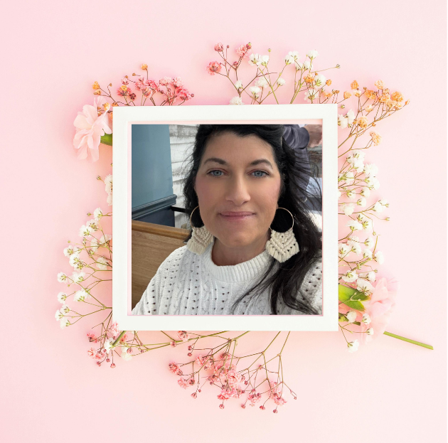 A woman with dark hair and blue eyes, wearing a white knitted sweater and large hoop earrings, taking a selfie indoors with a blurred background. The photo is framed with white borders and surrounded by pink and white baby's breath flowers on a pink background.