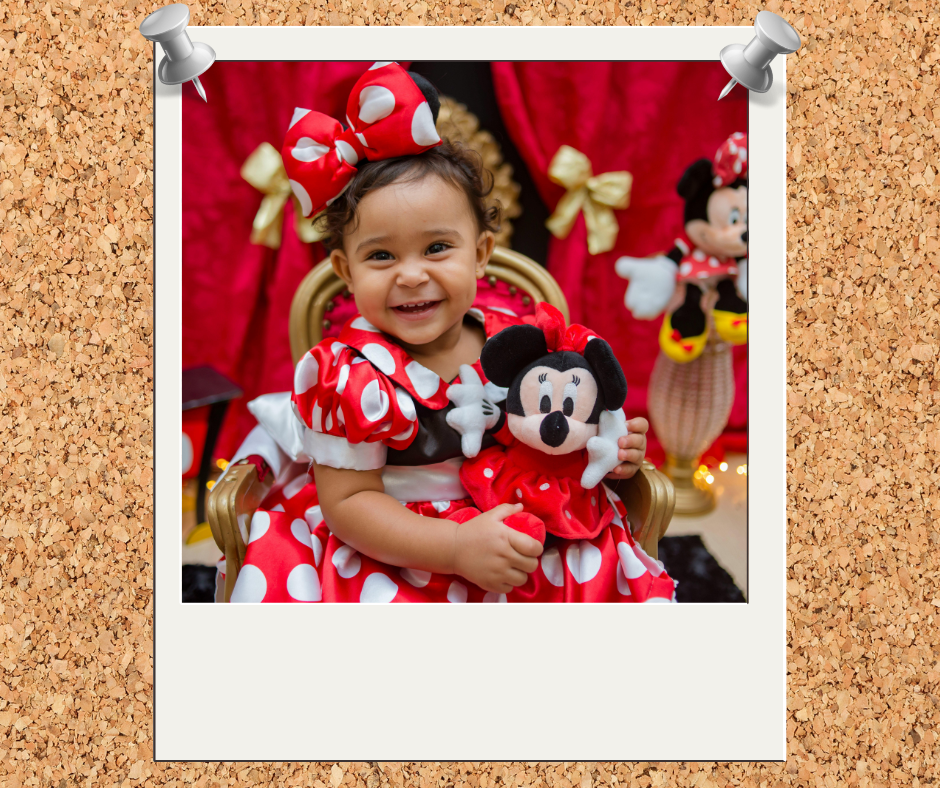 A smiling young girl with curly hair wearing a red and white polka dot dress and matching bow, holding a Mickey Mouse plush toy, with Minnie Mouse decorations in the background.