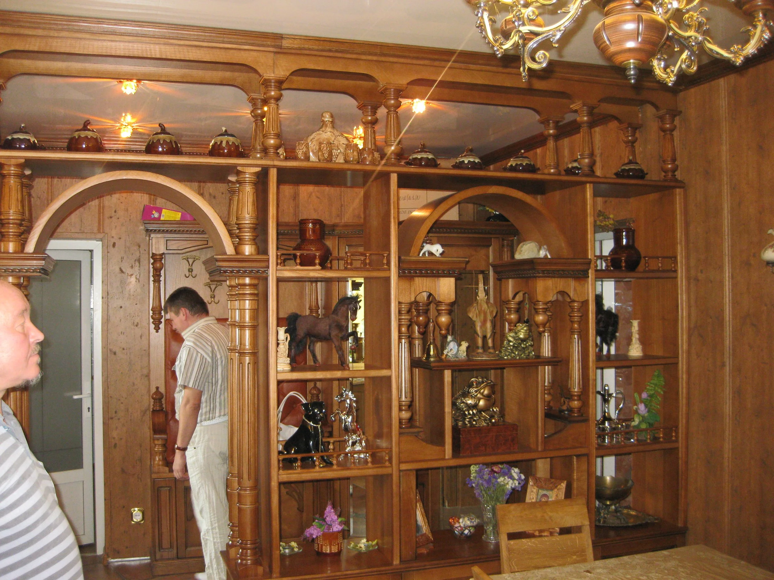 Interior of a wood-paneled room with a large wooden decorative shelving unit displaying various decorative items including vases, figurines, and flowers. Two men are present, one on the left side, partially visible, the other in the center, looking d