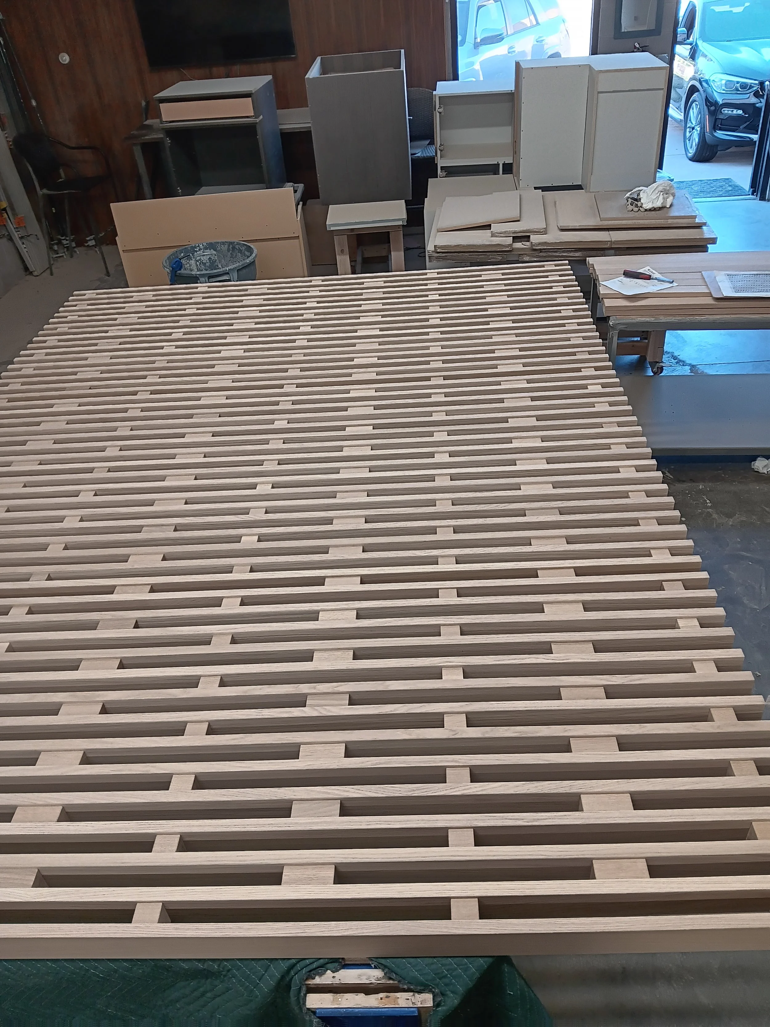 A woodworking workshop with a large slatted wooden tabletop in the foreground and various woodworking projects and tools in the background.