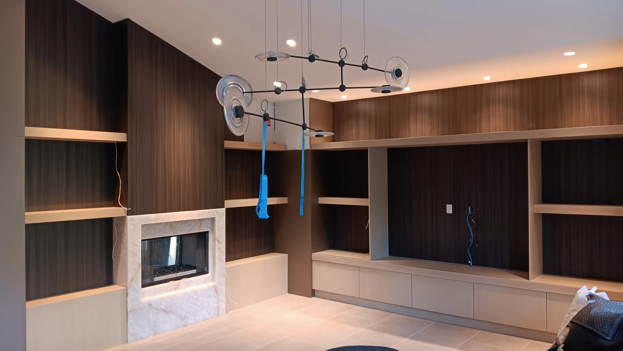 Interior view of a modern living room with dark wood paneled walls, built-in shelving, a fireplace with a marble surround, and a ceiling light fixture with multiple glass discs.