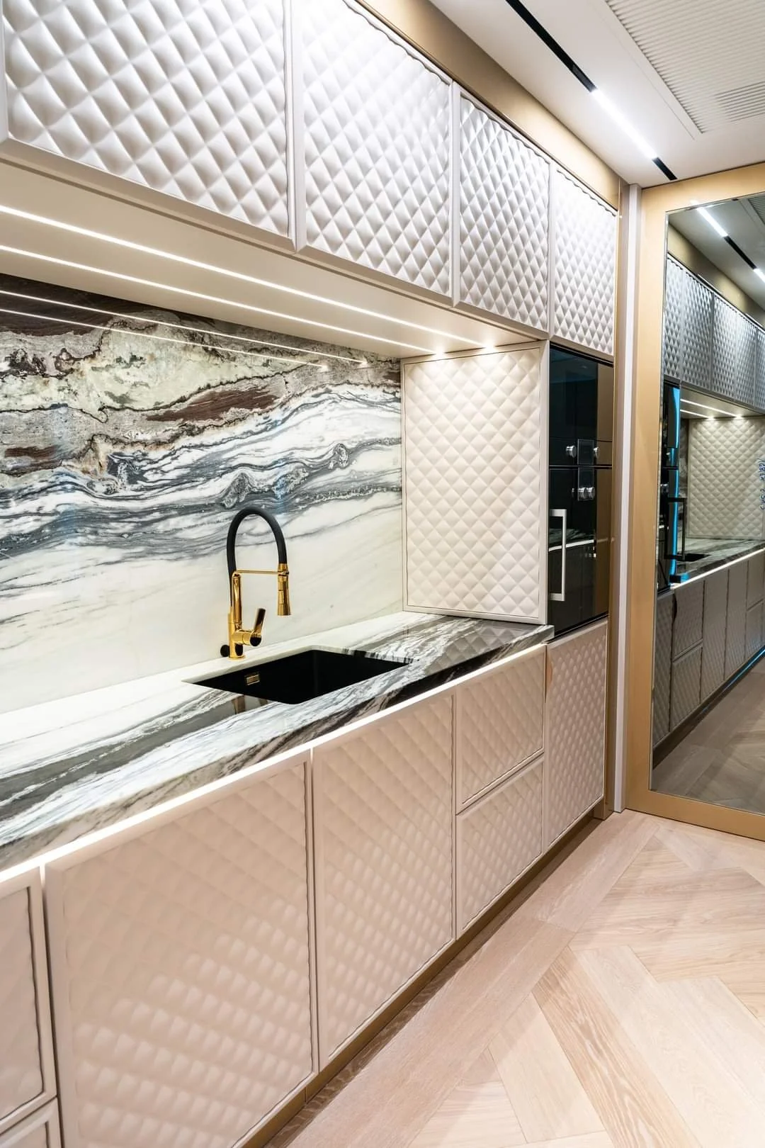 Modern kitchen with cream quilted cabinetry, a black sink, and gold faucet, featuring a marble backsplash with gray, white, and brown veins, and a reflective glass door with a reflection of the room.
