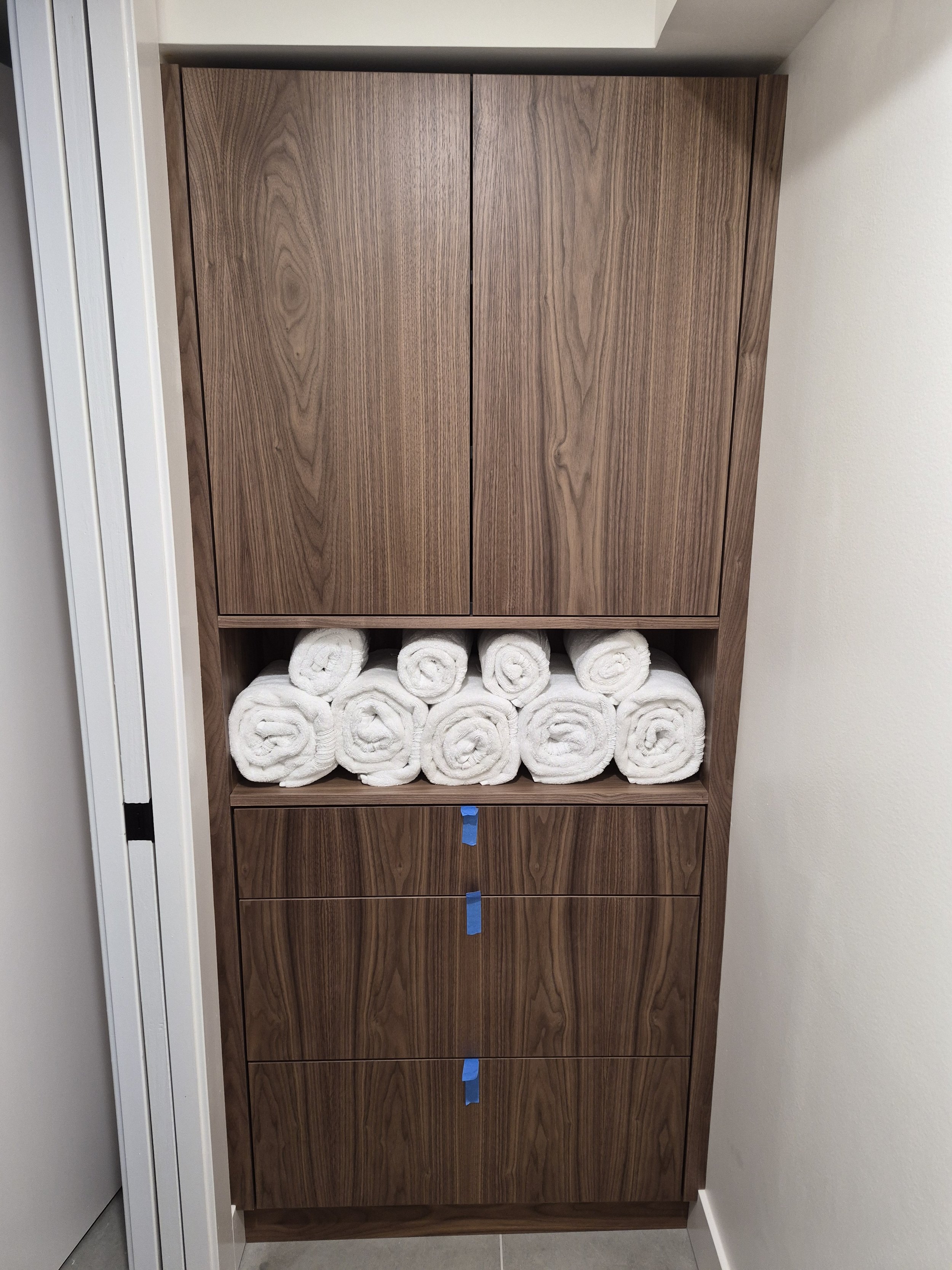 A wooden cabinet with closed doors at the top, an open shelf in the middle holding neatly rolled white towels, and three drawers at the bottom with blue painter's tape on each handle.