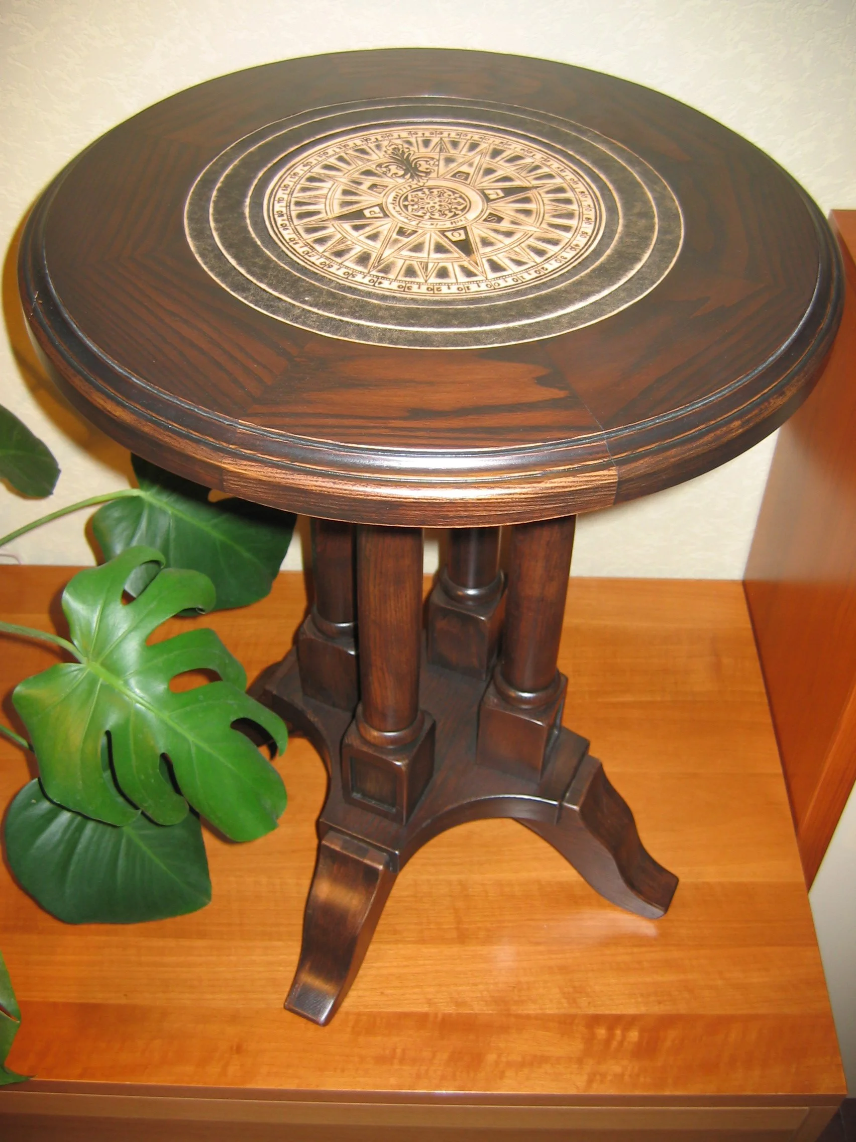 A round wooden table with an intricate compass design on the top surface, placed on a wooden floor, next to a green leafy plant.