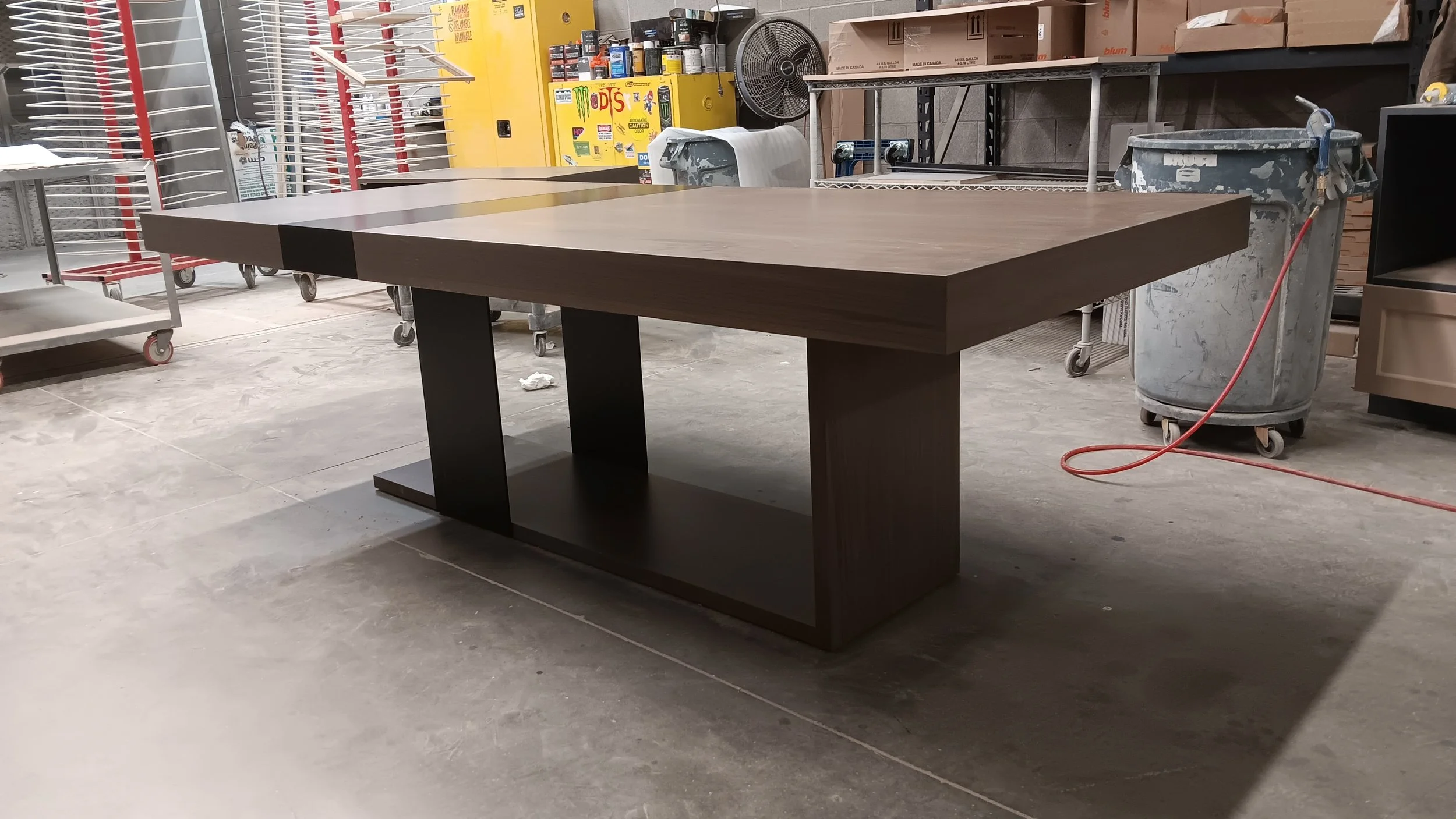 A modern wooden table with a dark brown finish, placed in an industrial workshop with shelves, carts, and equipment.