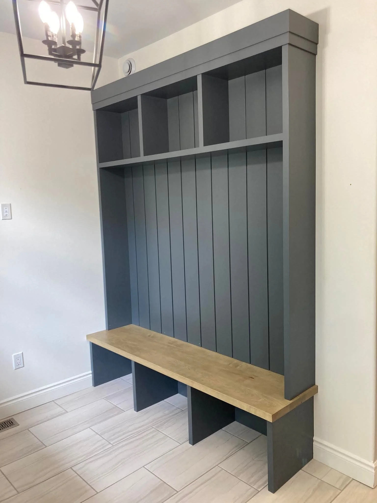 Built-in coat rack with cubbies, vertical slats, and a wooden bench underneath, in a hallway with beige tiled floor and white walls.