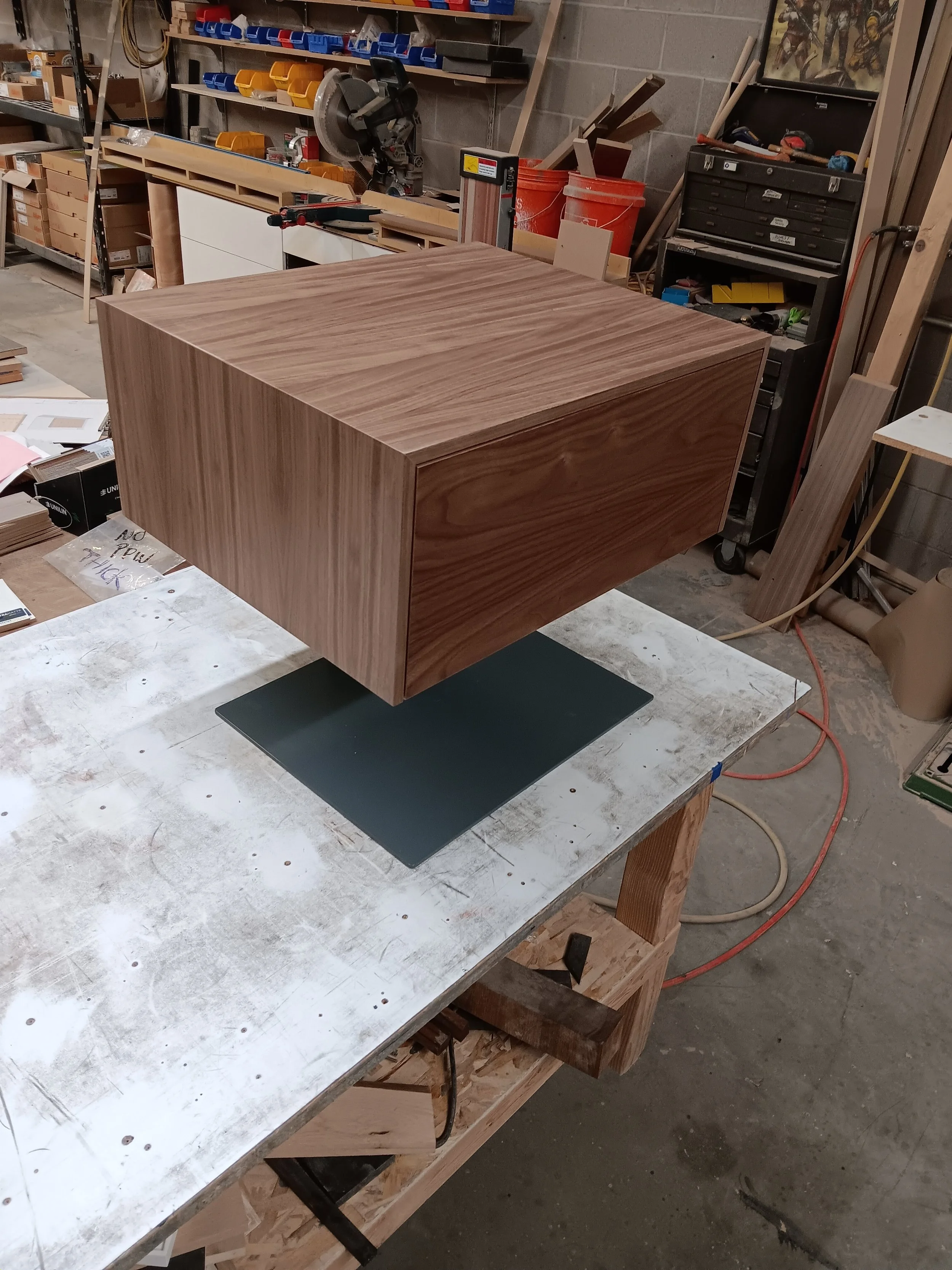 A wooden cube or box with a smooth surface and natural wood grain finish, placed on a black square base on a workbench in a woodworking workshop.