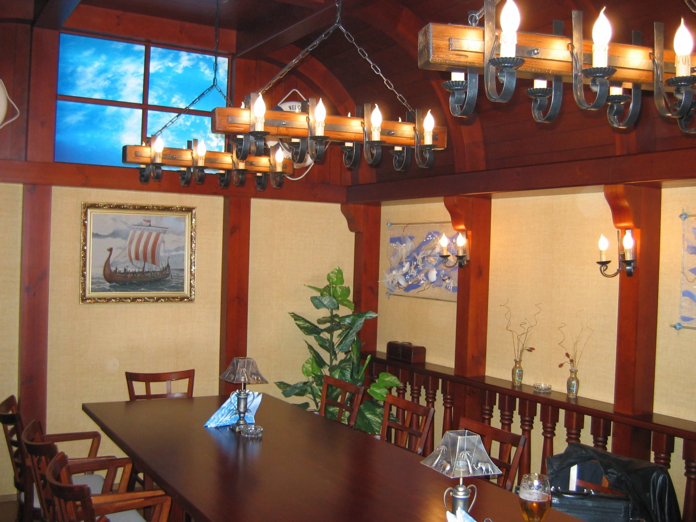 A dining room with wooden walls, a long dining table, and chairs. There are hanging chandeliers with candle-like lights and wall-mounted lights. A painting of a Viking ship is on the wall, and a window shows a blue sky with clouds.