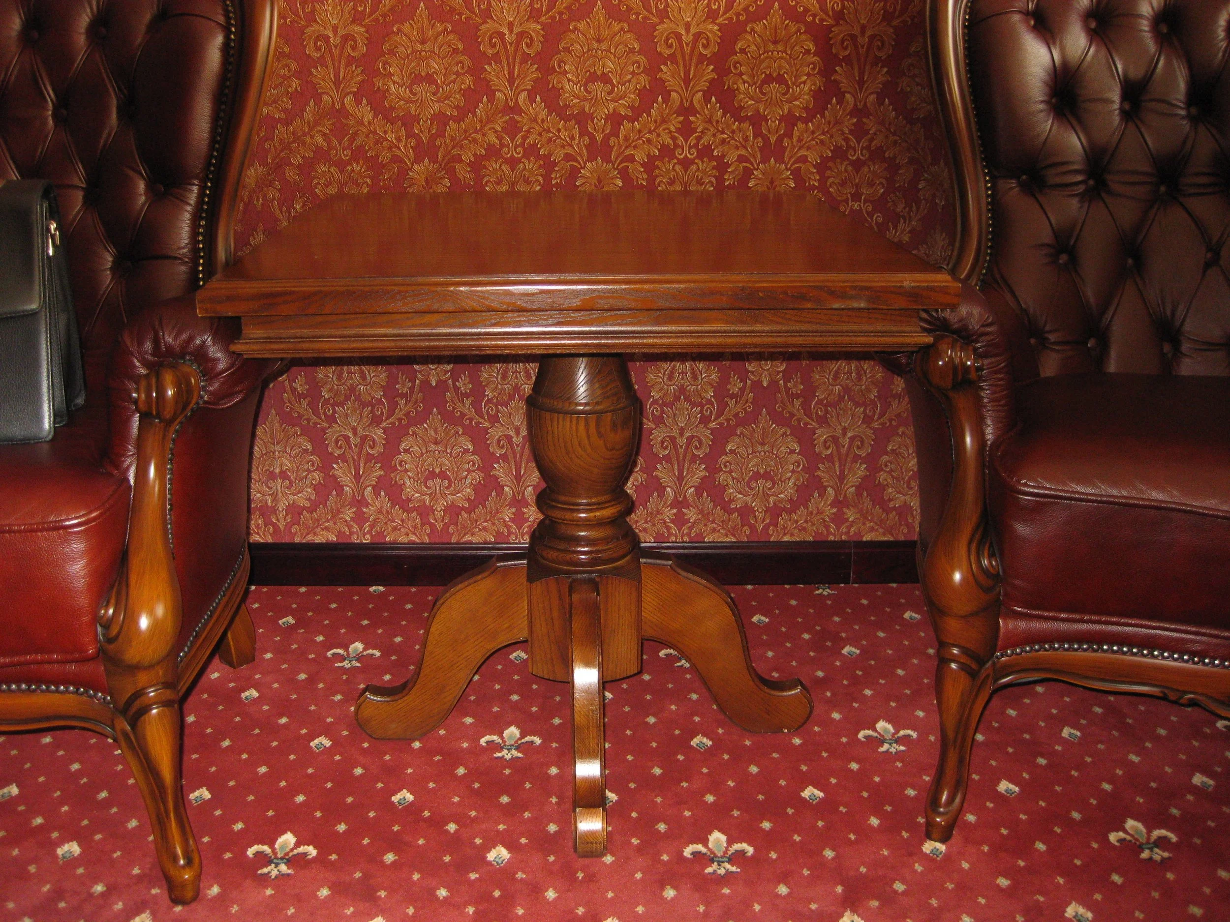 A wooden table with a pedestal base situated between two leather armchairs in a room with ornate red and gold wallpaper and a red patterned carpet.