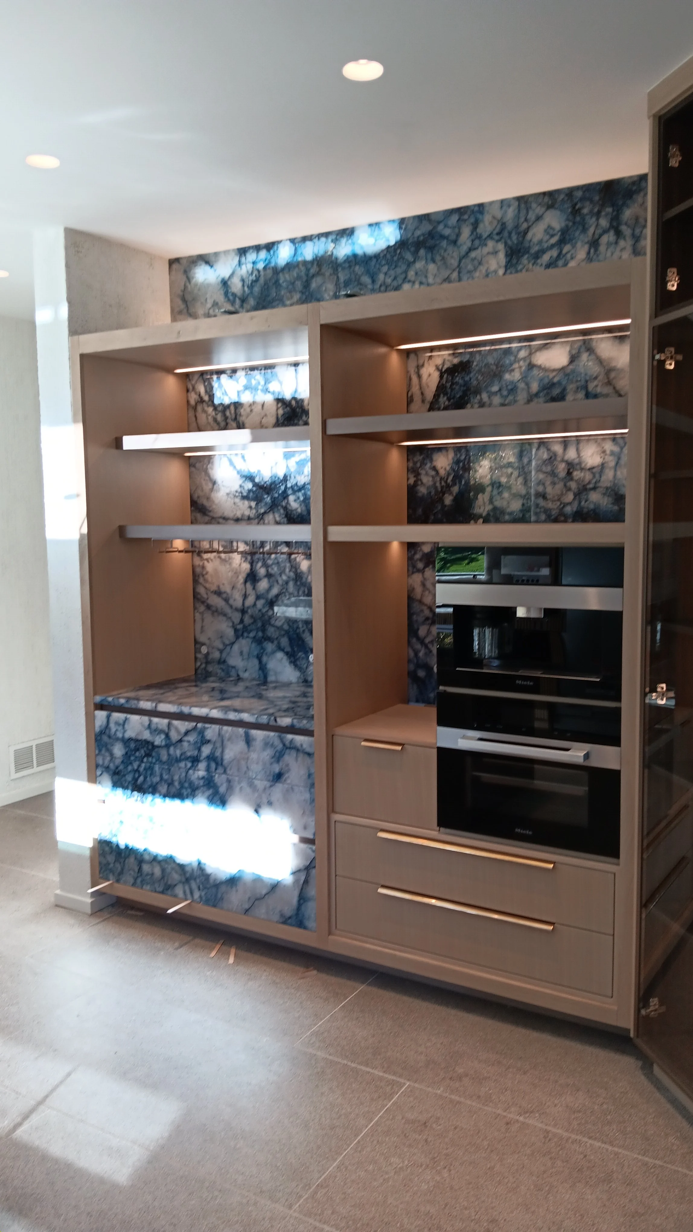 Modern built-in wooden cabinets with blue and white marble backsplashes and shelves, containing built-in appliances, in a well-lit room.