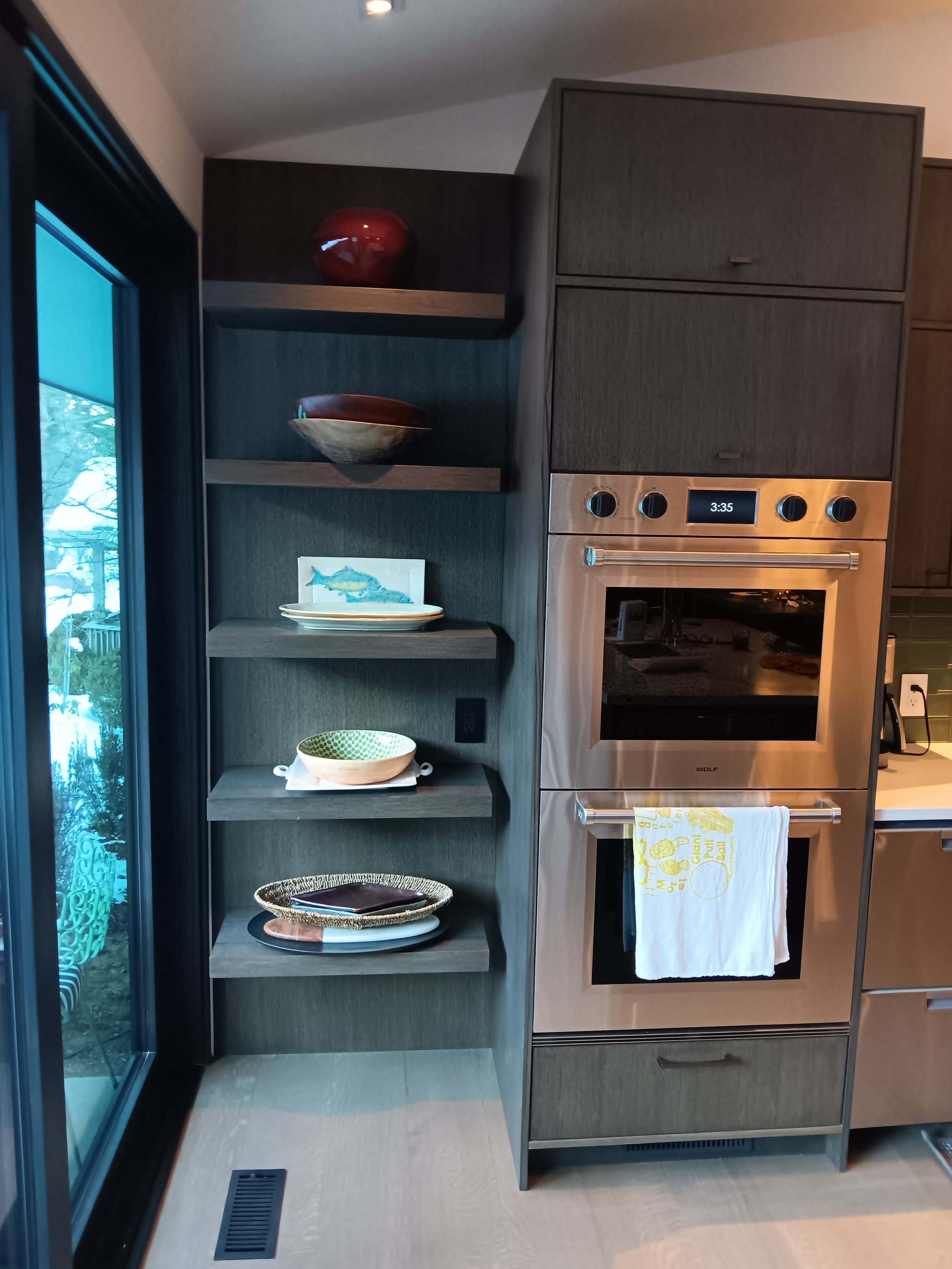 Kitchen corner with dark wood shelving and built-in oven, decorated with colorful bowls, plates, and a small fish illustration, near a sliding glass door.
